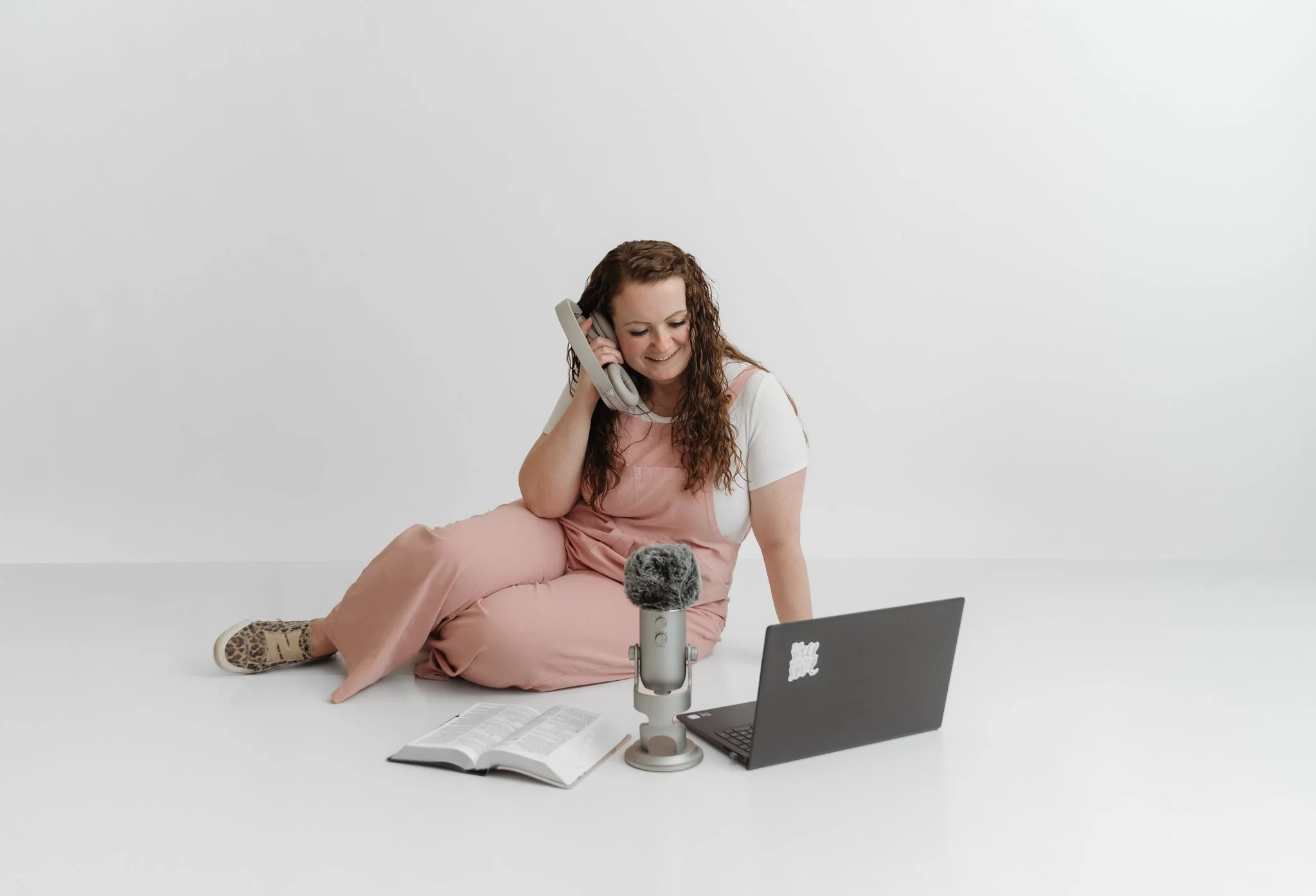A woman with long curly hair sits on the floor in a white room, smiling while talking into a gray microphone. She is wearing a casual pink jumpsuit with a white shirt underneath and leopard-print shoes. A laptop, an open book, and a sheet of paper are on the floor in front of her. She is holding a gray phone to her ear as she looks down.