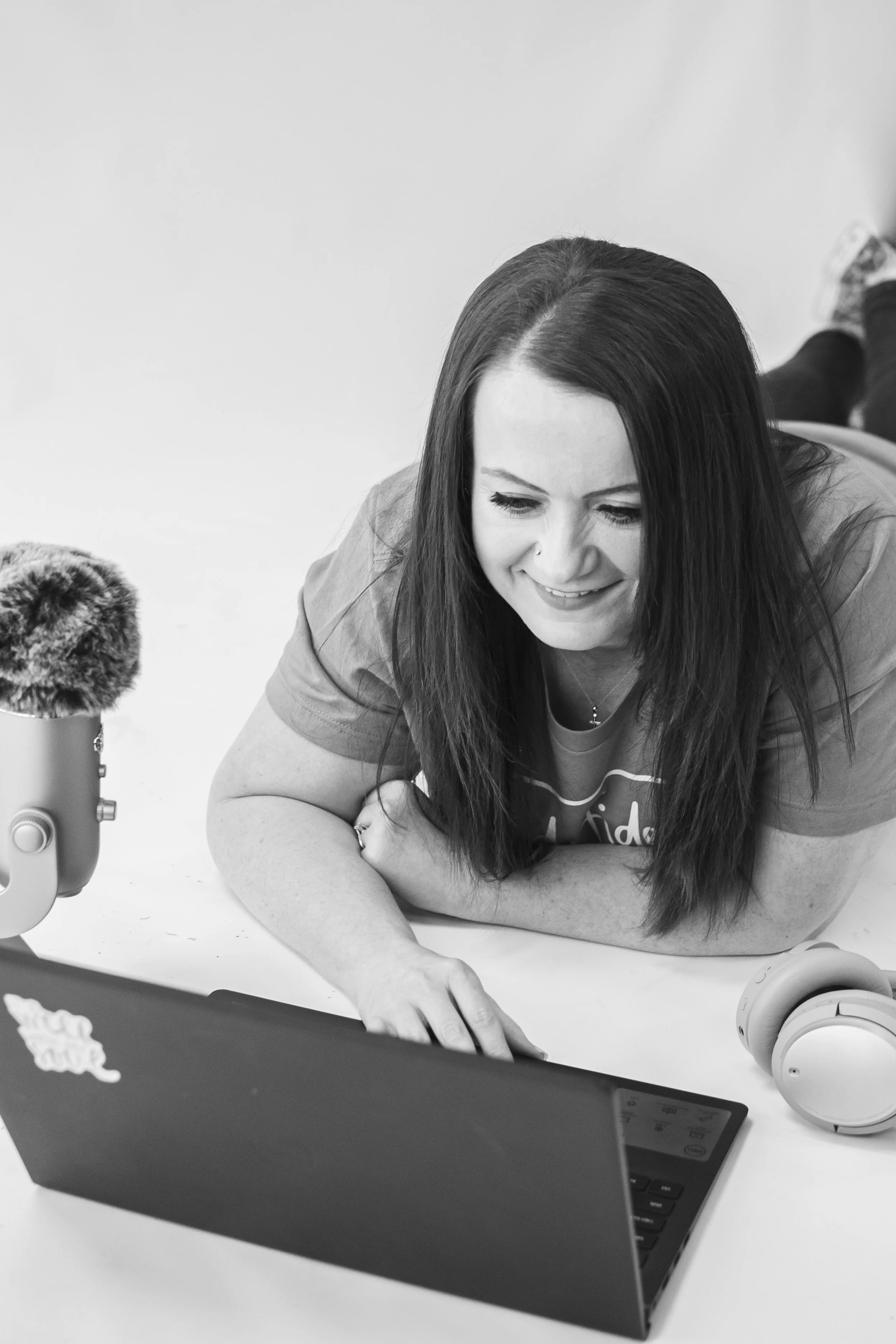 A woman with long dark hair, lying on her stomach, smiling and looking at a laptop screen. She has a nose piercing and is wearing a T-shirt. There's a microphone with a furry windscreen and a pair of headphones on the table next to her.