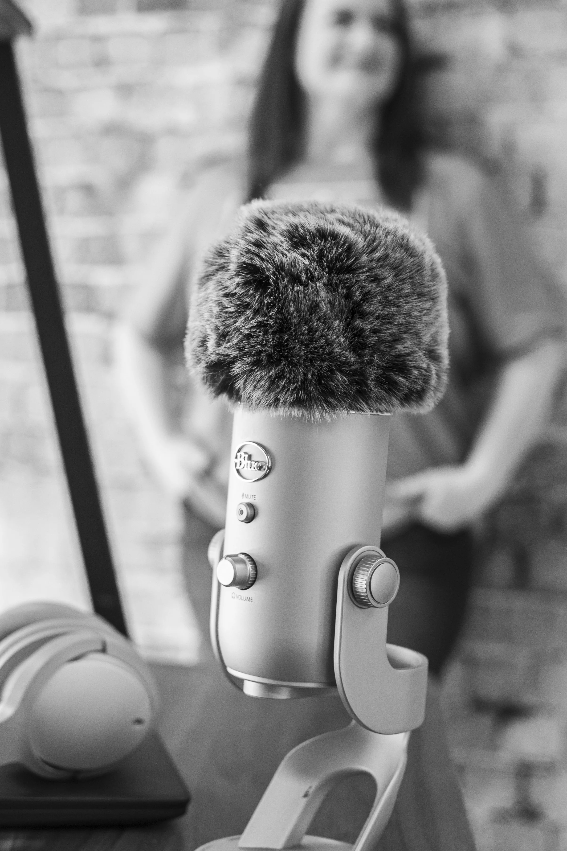 A close-up of a Blue Yeti microphone with a furry windscreen, set on a desk with headphones nearby. In the blurred background, a woman is sitting in front of a brick wall.