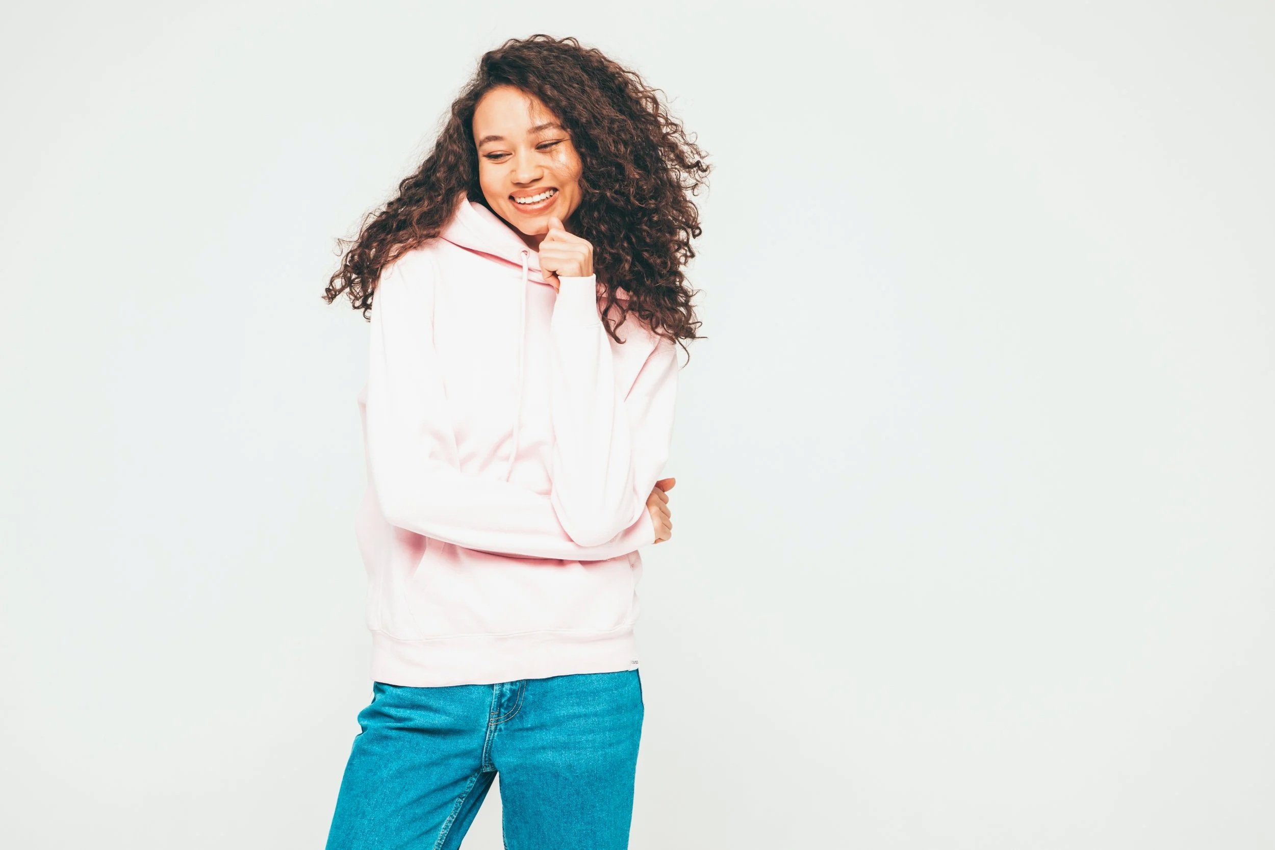 A teen with curly hair smiling and wearing a pink hoodie and blue jeans, standing against a plain grey background.