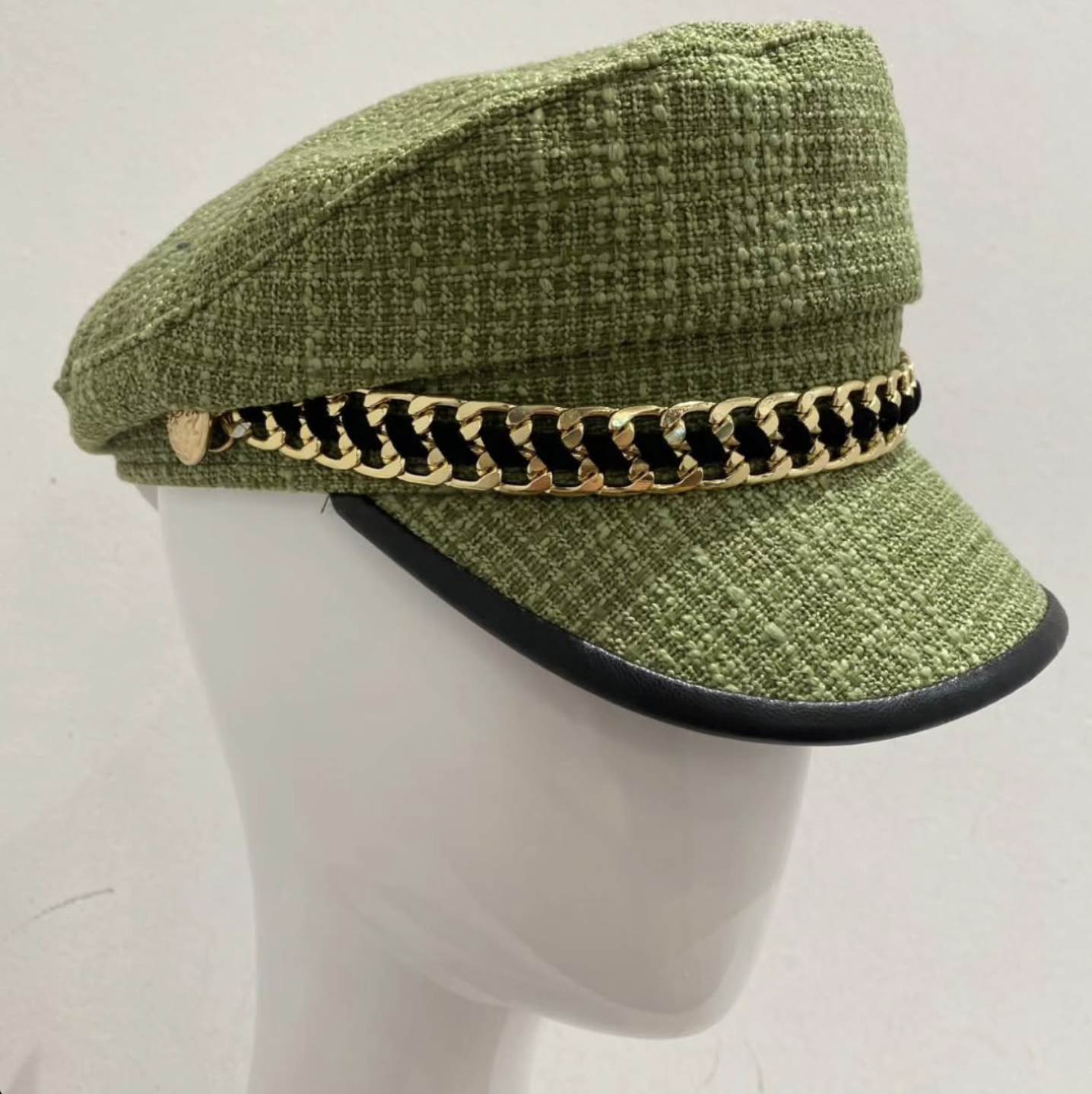 Green textured fabric cap with a decorative black and gold chain band and a small gold emblem, displayed on a white mannequin head.