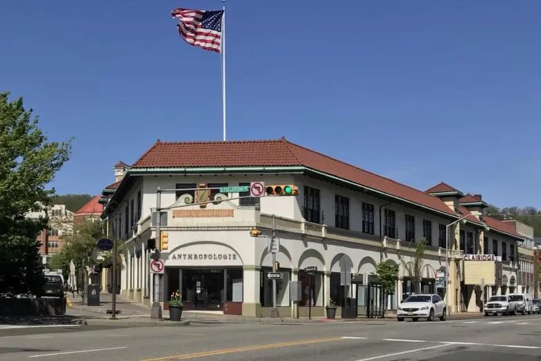 A corner building with a large American flag on a pole on top, featuring a red-tiled roof, multiple windows, and several signs including 'ANTHROPOLOGIE' and 'CLAIRIDGE,' located in an urban area with trees, cars, and street signs.