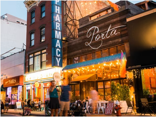 Night view of a street with shops and restaurants, illuminated by string lights, with people walking and sitting outside.