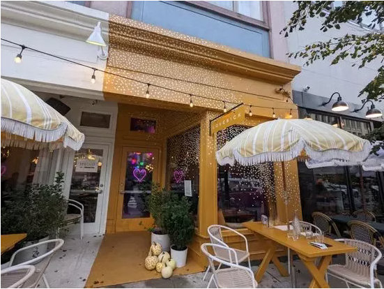 Outdoor patio of a cafe with umbrellas, outdoor tables, and string lights, located in an urban area with buildings and trees.