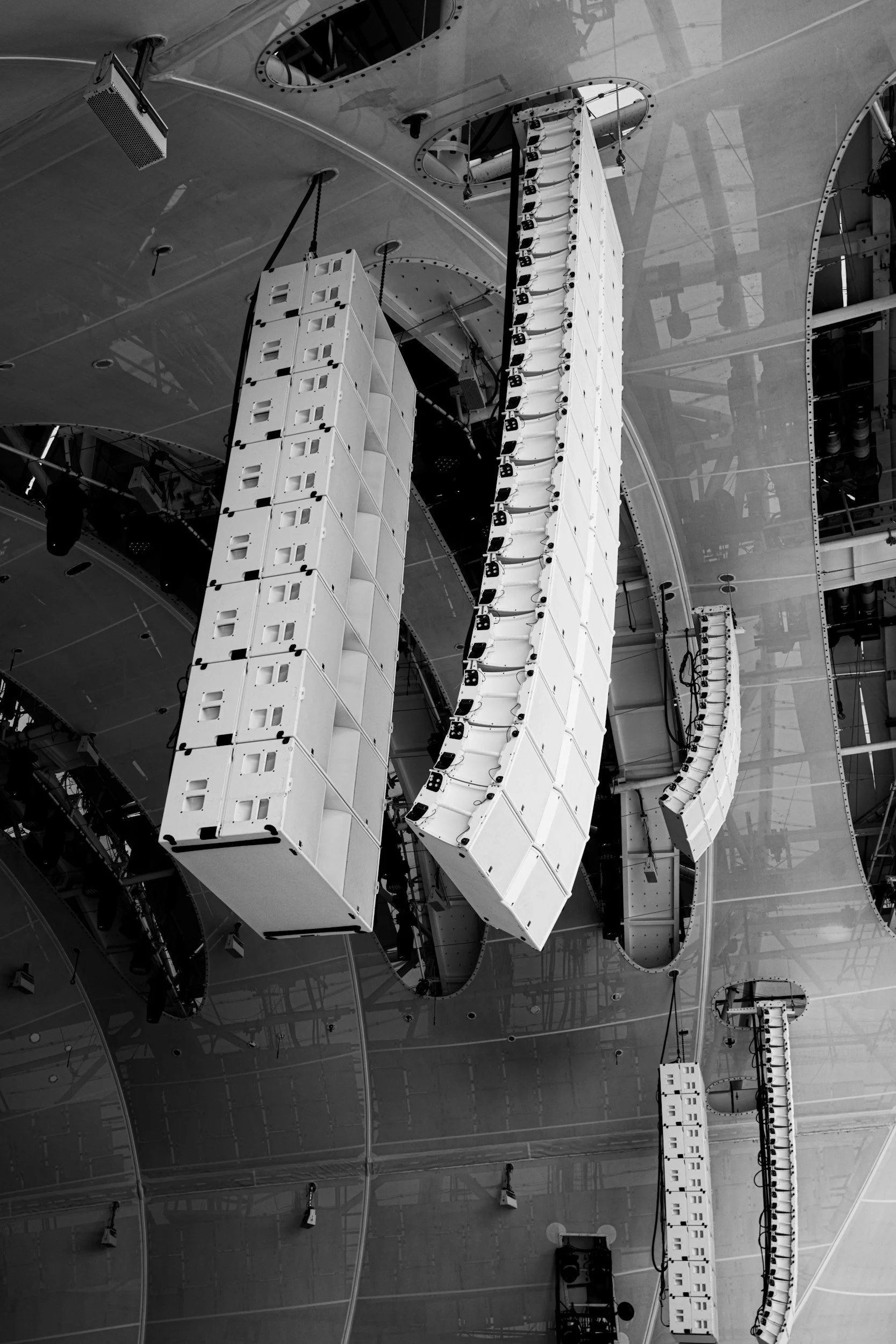 Black and white photo of large speaker arrays hanging from a ceiling at a concert or event venue.
