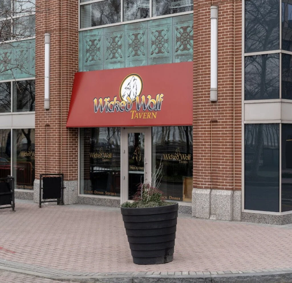 The exterior of Wicked Wolf Tavern, a brick building with a red awning displaying the tavern's name and logo, a white wolf head, underneath a window with decorative panels.