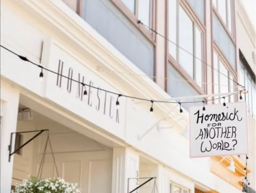 A sign on a building reads "Homesick for another world?" next to a string of lights that spell out "Holostia." The building has large windows and a decorative facade.