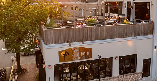 The image shows a two-story building with a restaurant on the second floor, with outdoor seating on a rooftop patio. There are people dining at the outdoor tables, and a large tree visible to the left of the building.