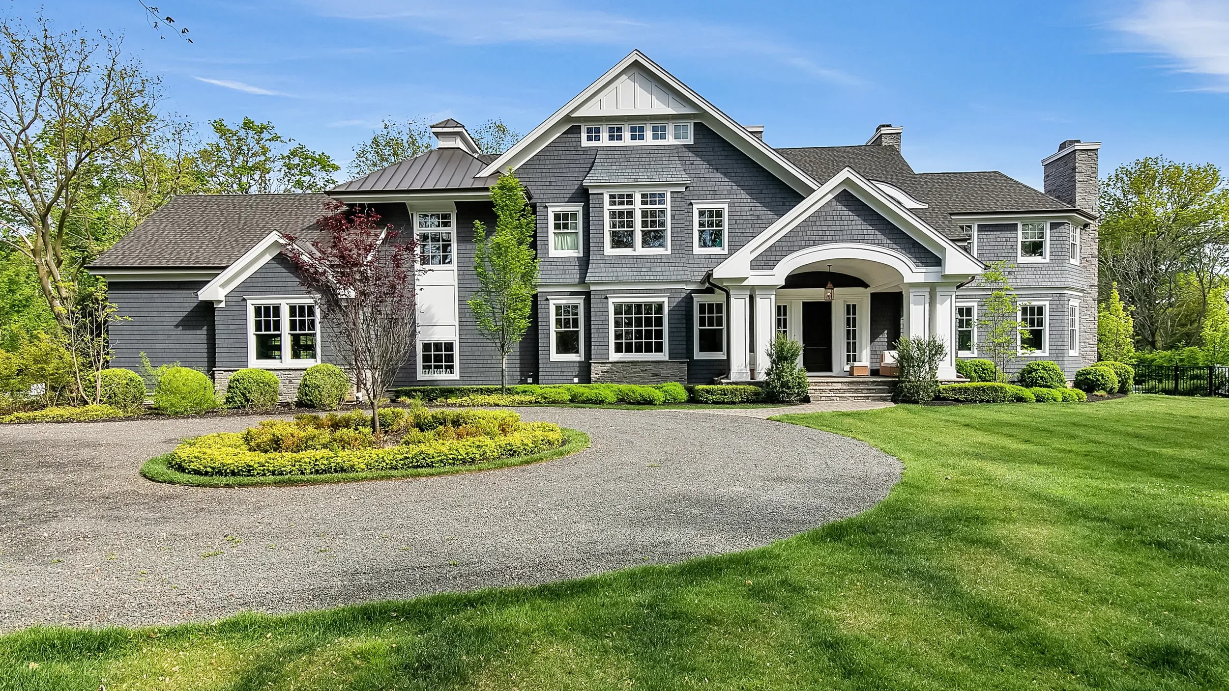 A large, modern gray house with white trim, surrounded by a well-maintained lawn and trees, with a gravel driveway leading to the front porch and entrance.