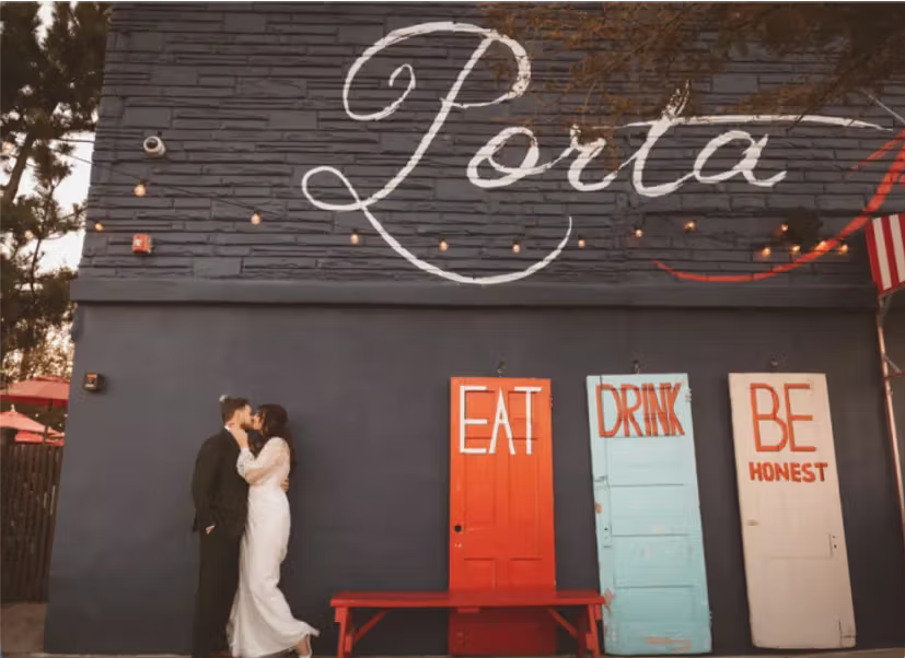 A couple dressed in wedding attire kissing in front of a dark-colored wall with the word 'Porta' painted on it. Below, there are three colorful signs that say 'EAT,' 'DRINK,' and 'BE HONEST.'