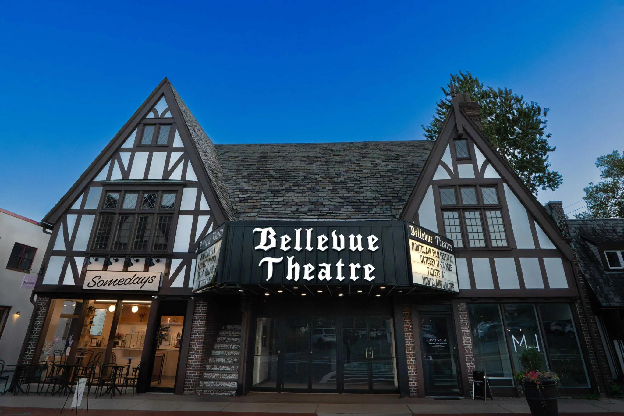 The Bellevue Theatre building with a Tudor Revival style, featuring a black marquee with white illuminated letters, a sign for 'Somedays' restaurant, and a sidewalk seating area at dusk.
