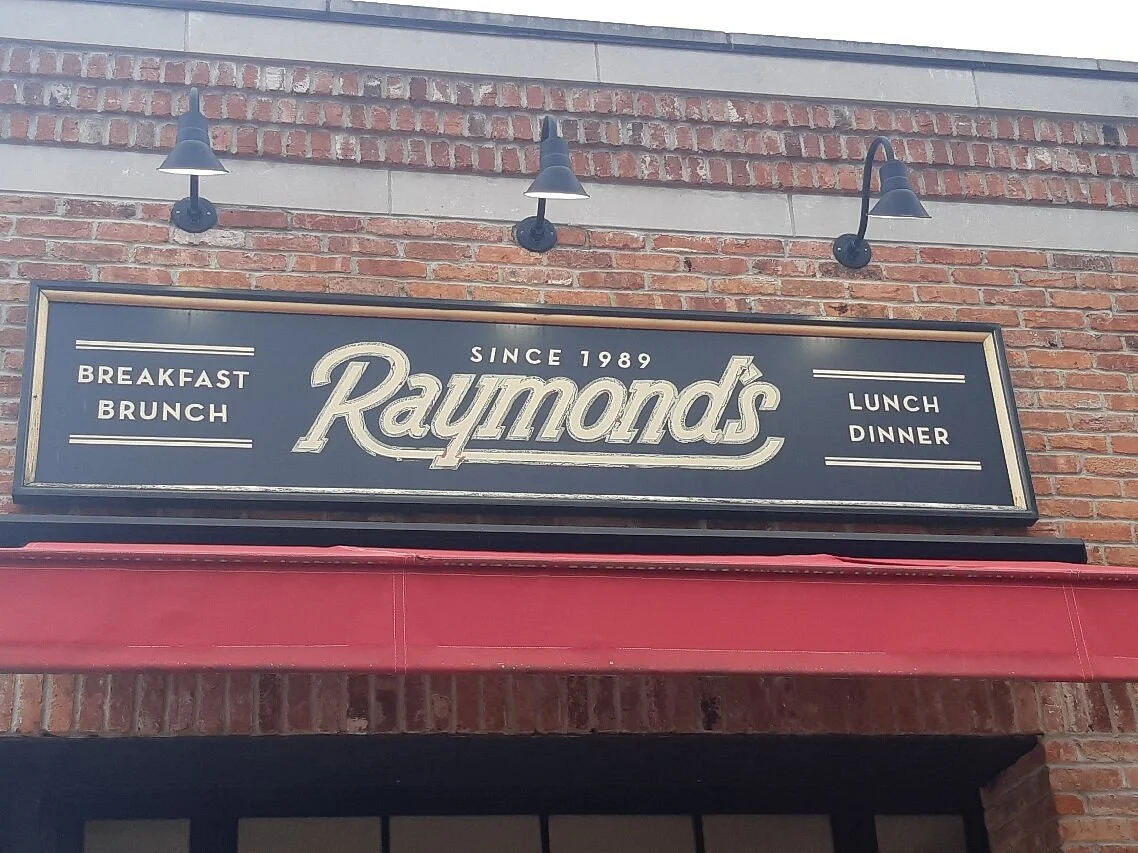 Exterior sign for Raymonds restaurant on brick building, offering breakfast, brunch, lunch, and dinner, established in 1989.