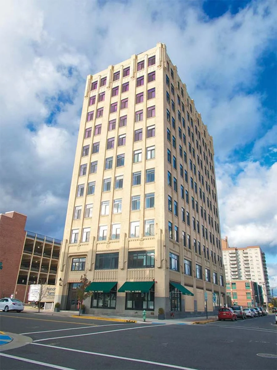 A tall beige building with multiple windows and green awnings at the ground level, set against a bright blue sky with scattered clouds, in an urban area with parked cars and other buildings.