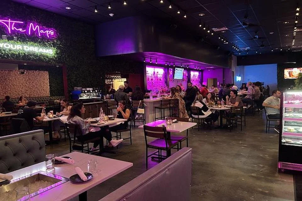 People dining in a modern restaurant with purple and pink lighting, dark walls, and a bar area with TVs.