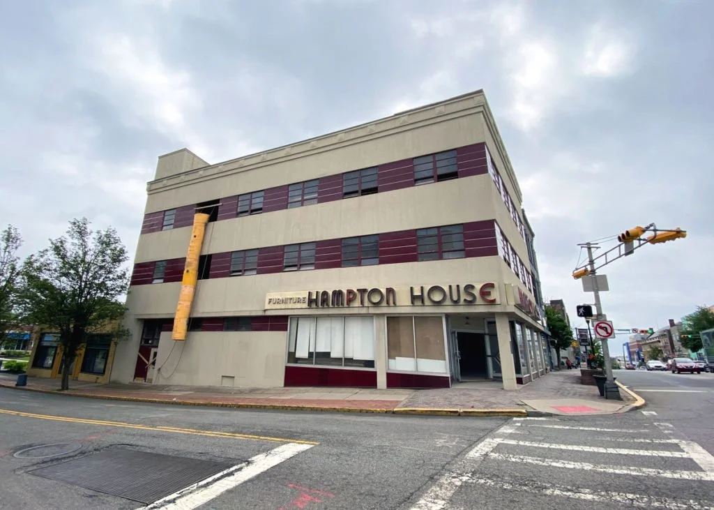 Street view of a corner building labeled 'HAMPTON HOUSE' with a sign for furniture. The building has horizontal burgundy stripes and windows with dark frames. A yellow pipe runs down the side of the building. There are trees, street crossings, and traffic signals visible, with cloudy skies overhead.