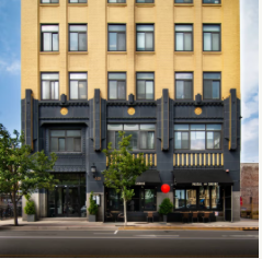 Facade of a multi-story building with a black and yellow exterior, windows, trees, and a sidewalk in front.