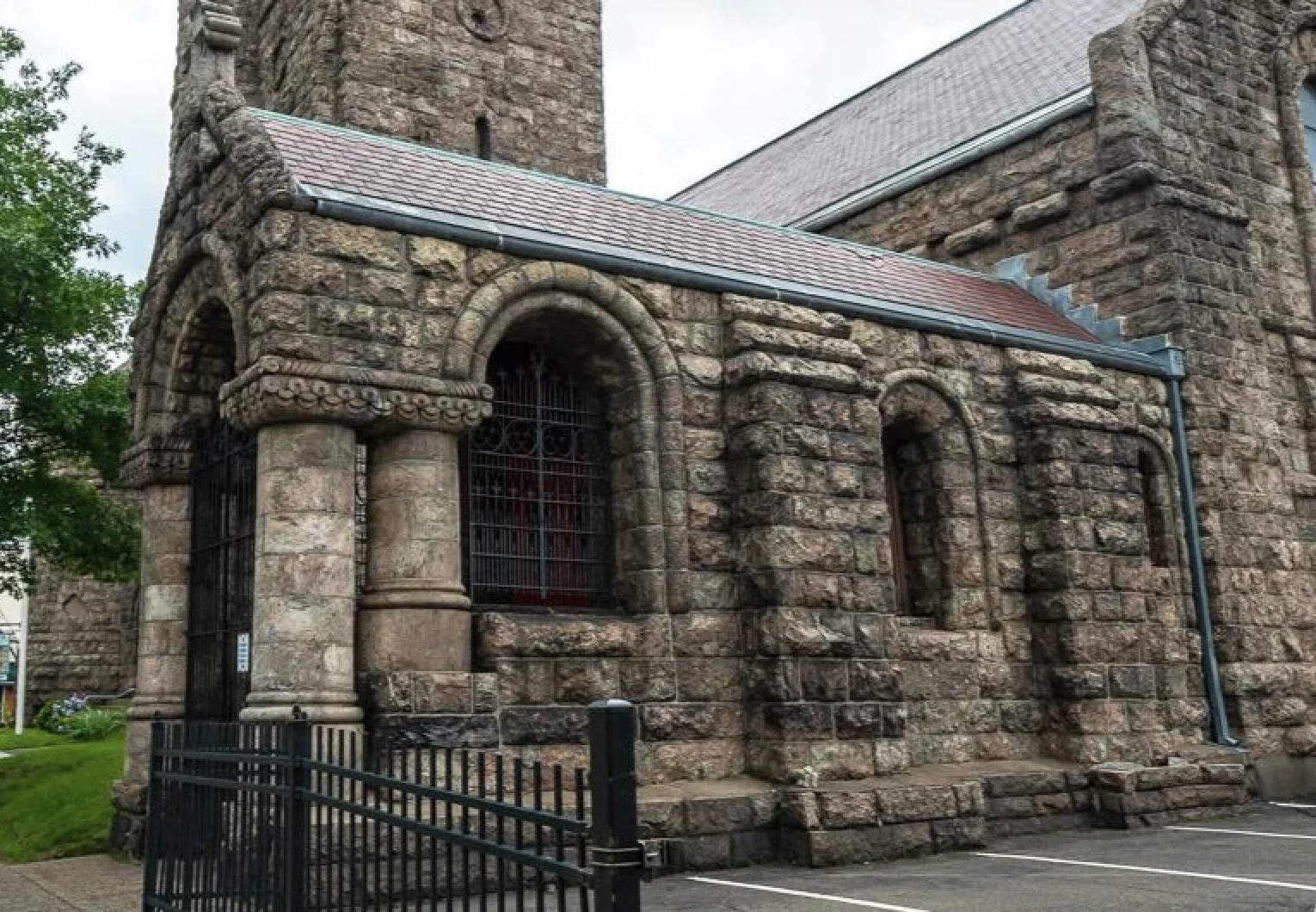 A stone church building with arched windows and a steep roof, surrounded by a black metal fence.