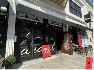Exterior of a storefront with large glass windows, an 'OPEN' sign on the sidewalk, and outdoor seating with tables and chairs.