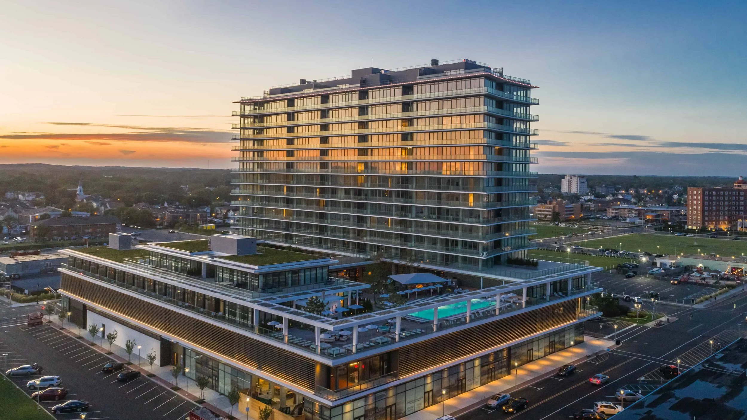 A modern multi-story building with glass balconies at sunset. The building has a rooftop pool and outdoor seating area, with a parking lot in the foreground and a cityscape background.