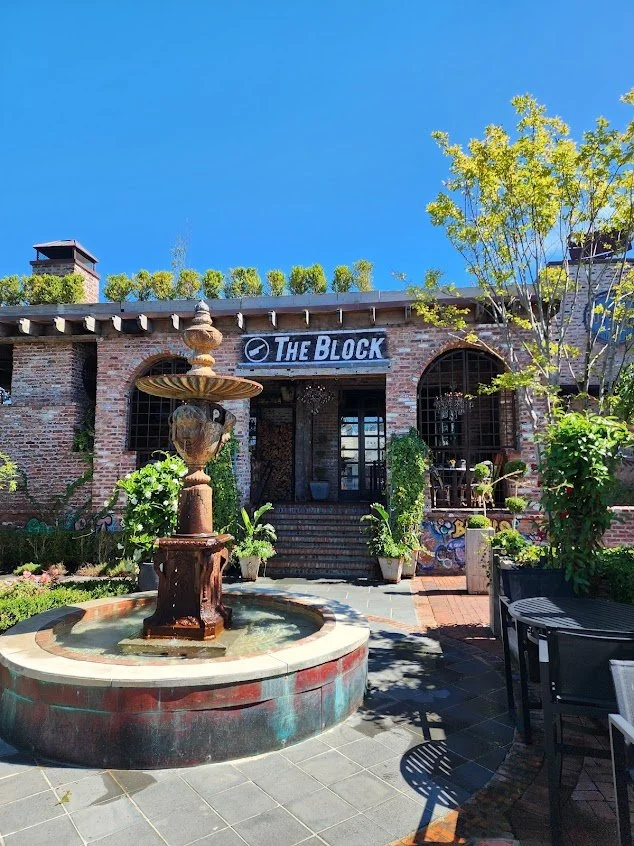 Exterior of a brick building with a fountain in front, surrounded by plants and outdoor seating, under a clear blue sky.