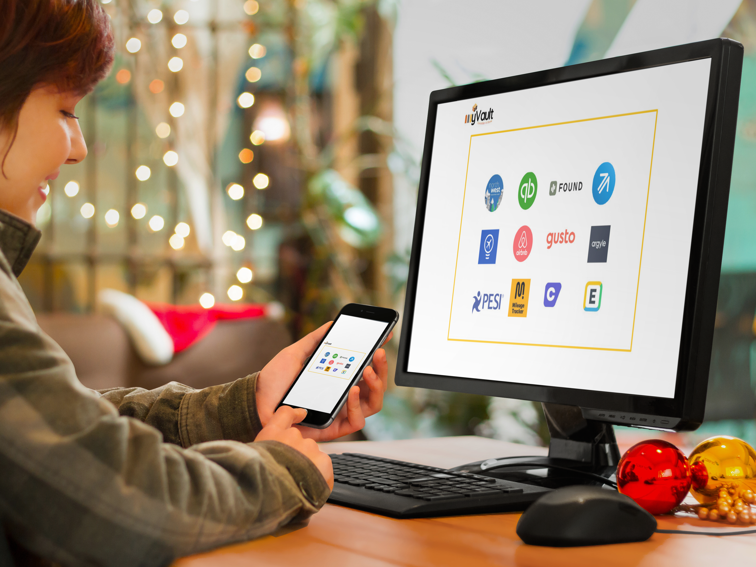 A woman looking at her smartphone with a Christmas-themed background, sitting at a desk with a computer monitor displaying various app logos, and decorated with red and gold ornaments.