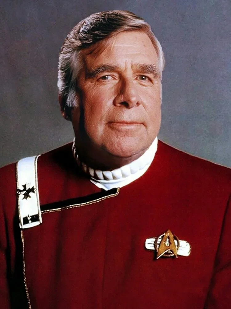 Star Trek creator Gene Rodenberry with gray hair wearing a red Star Trek uniform with a badge and a white sash, posing against a plain background.