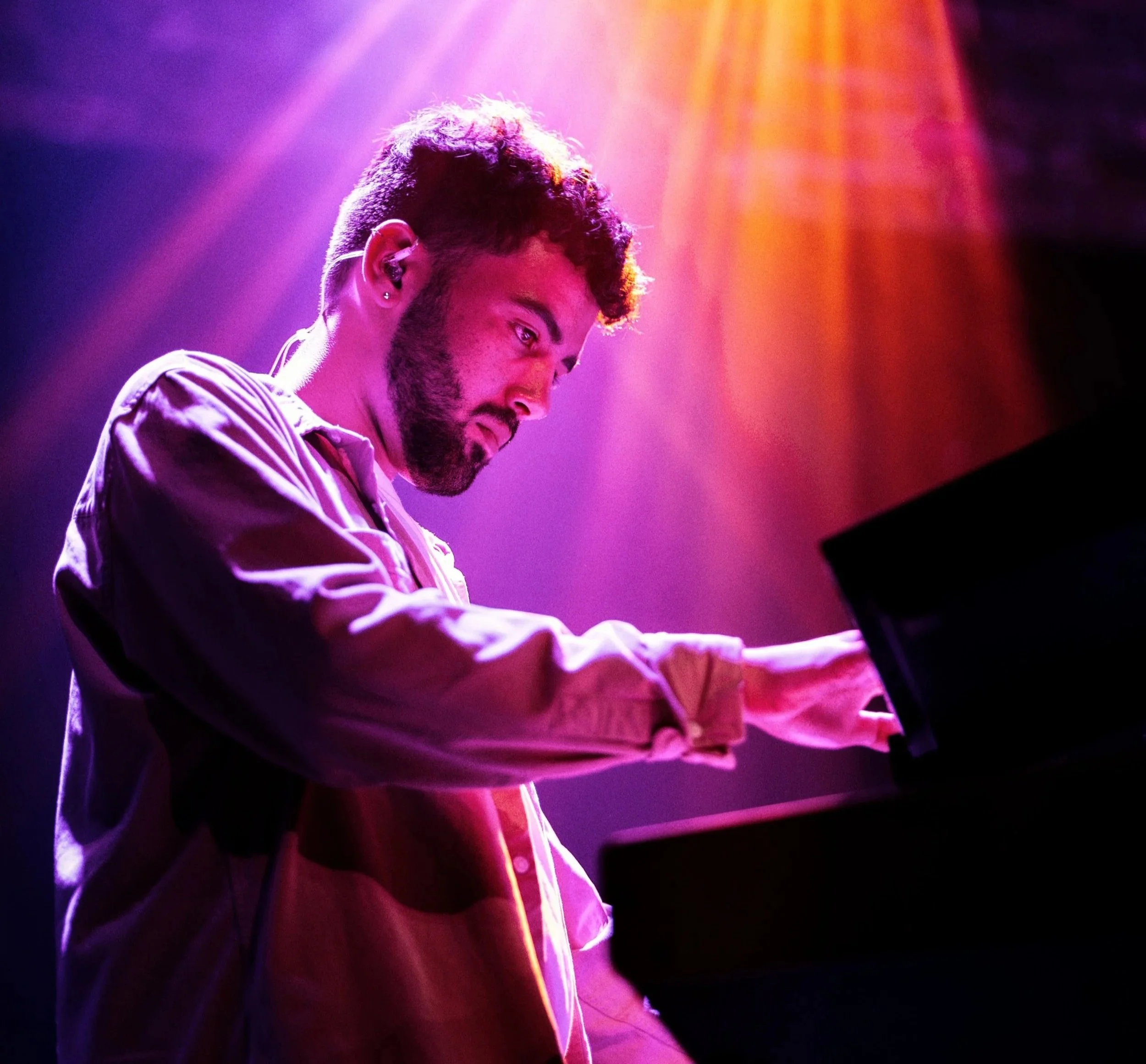 A man with dark hair and a beard playing a piano, lit by colorful stage lights in purple, pink, and orange hues.