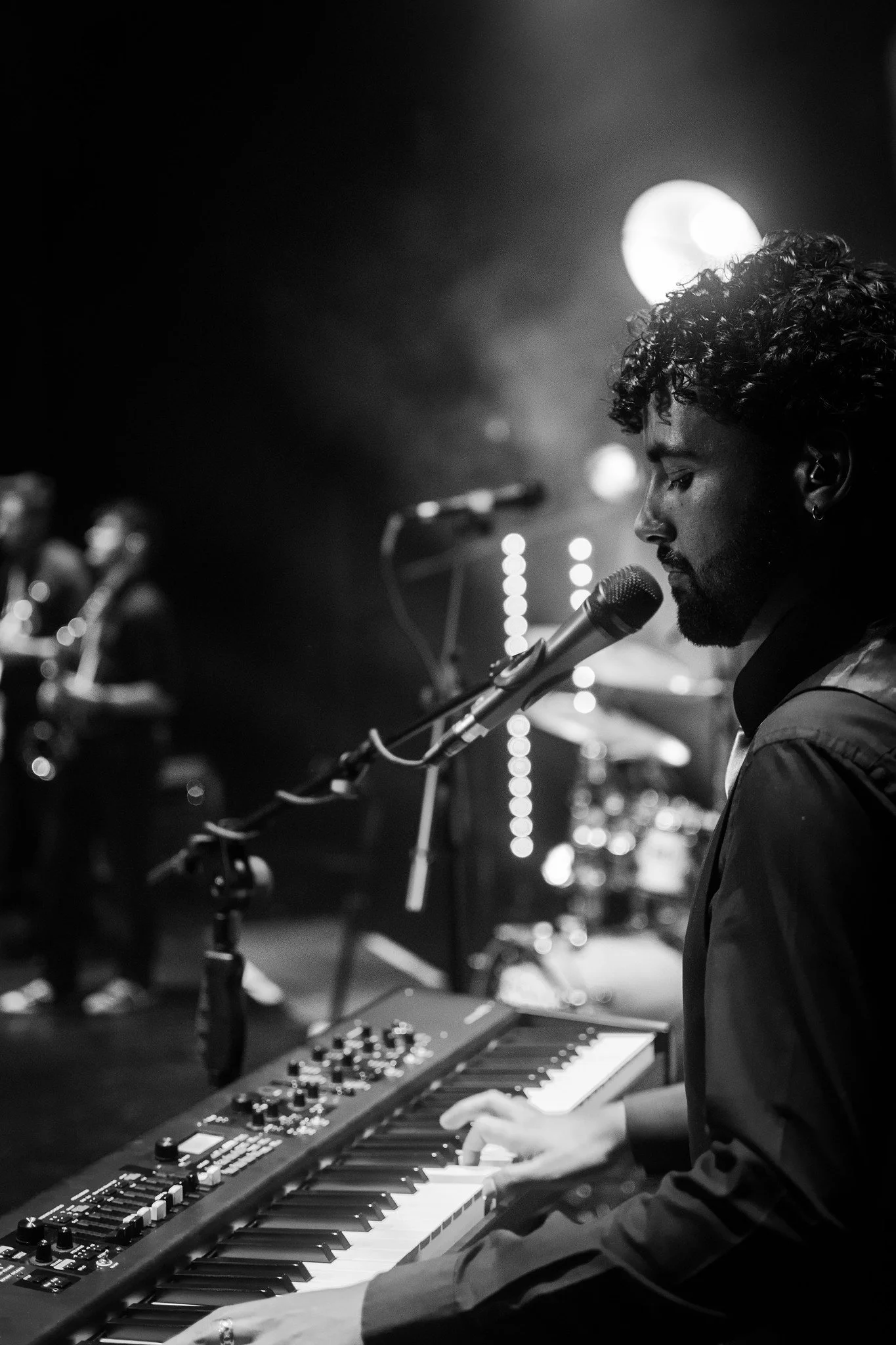Black and white photo of a musician with curly hair playing a keyboard with a microphone in front of him in a dimly lit setting.