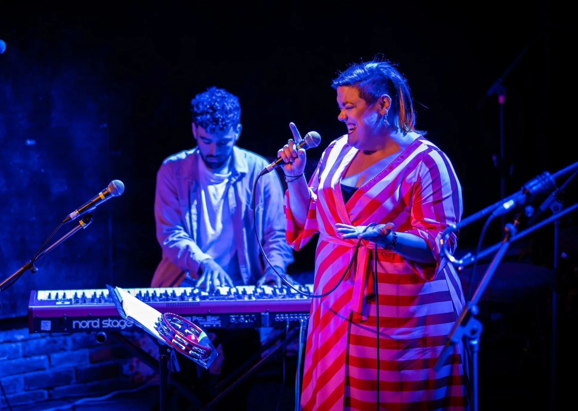 A female singer in a red and white striped outfit performs with a microphone, while a keyboard player in the background plays on stage with dark lighting.