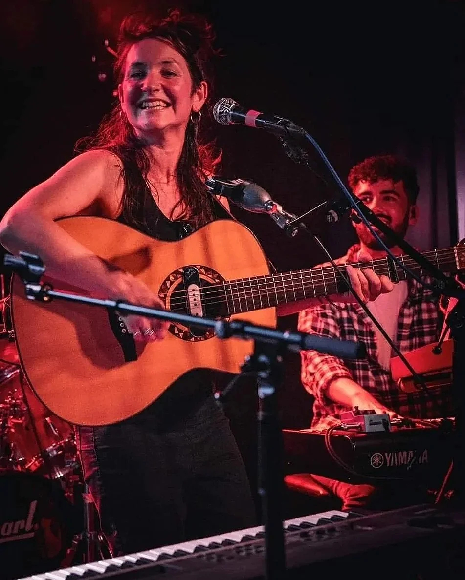 A woman playing an acoustic guitar and singing into a microphone on stage, smiling, with a man playing a keyboard behind her in a dimly lit setting.