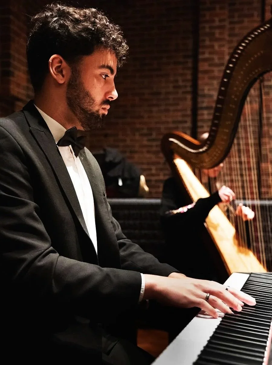 A man in a tuxedo playing a grand piano on stage with a brick wall background, while a person in black is playing a harp behind him.