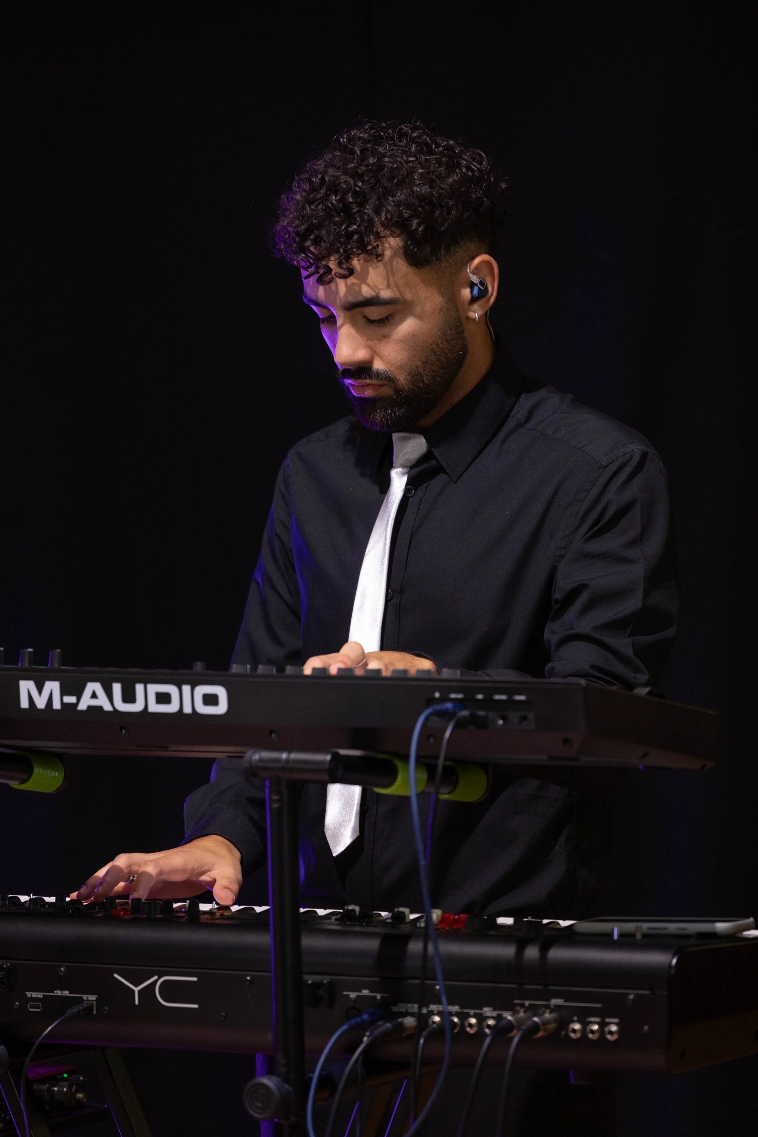 A male musician with dark curly hair and a beard playing a keyboard against a black background, wearing a black shirt, white tie, and in-ear monitors.
