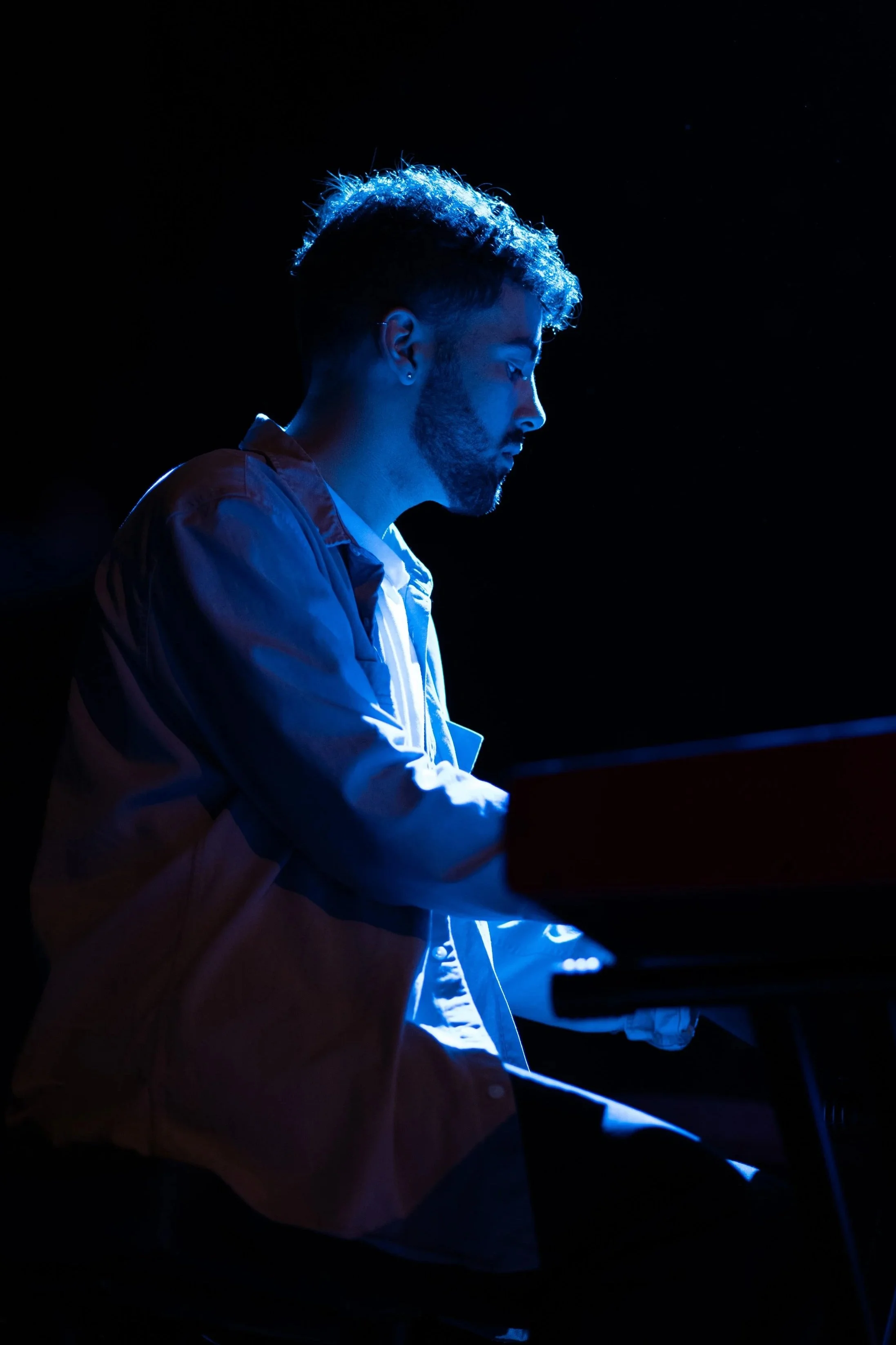 Side profile of a man with curly hair and beard playing the piano in a dimly lit room, illuminated by blue light.