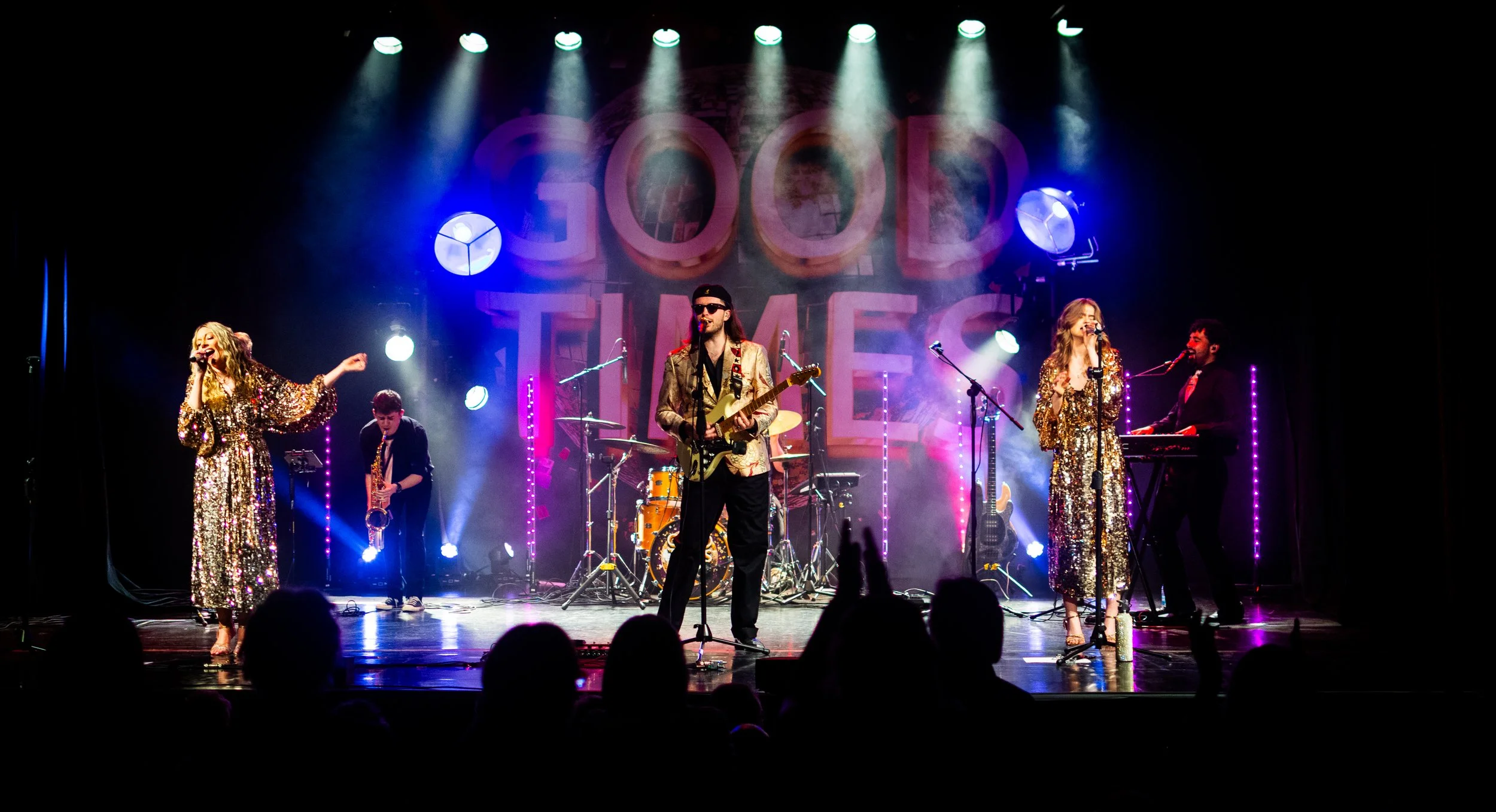 Band performing on stage with colorful lights, two women in gold dresses singing, a man with sunglasses playing guitar, and other musicians in the background.