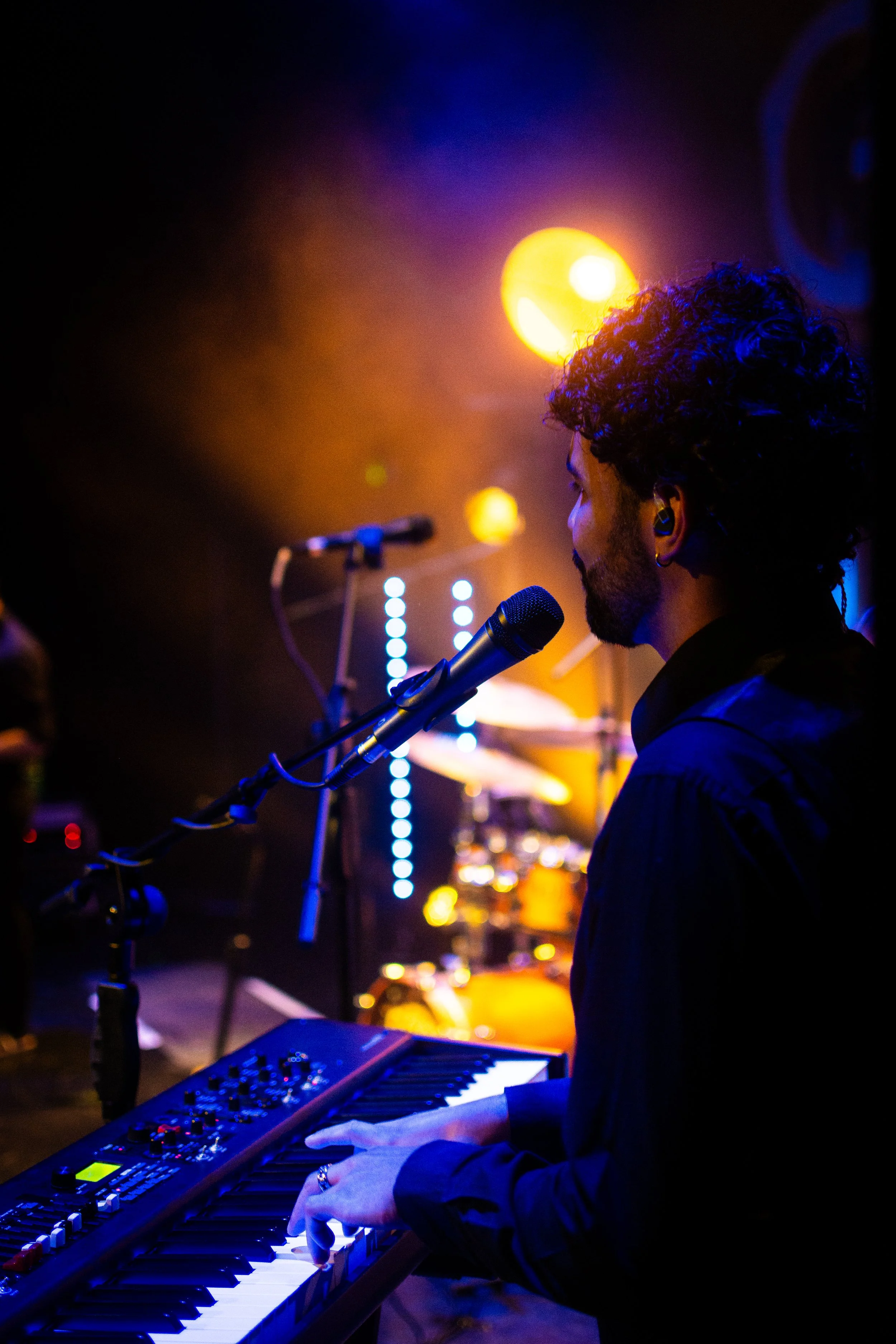 A person playing a keyboard on stage, with colorful stage lights and a dark background.