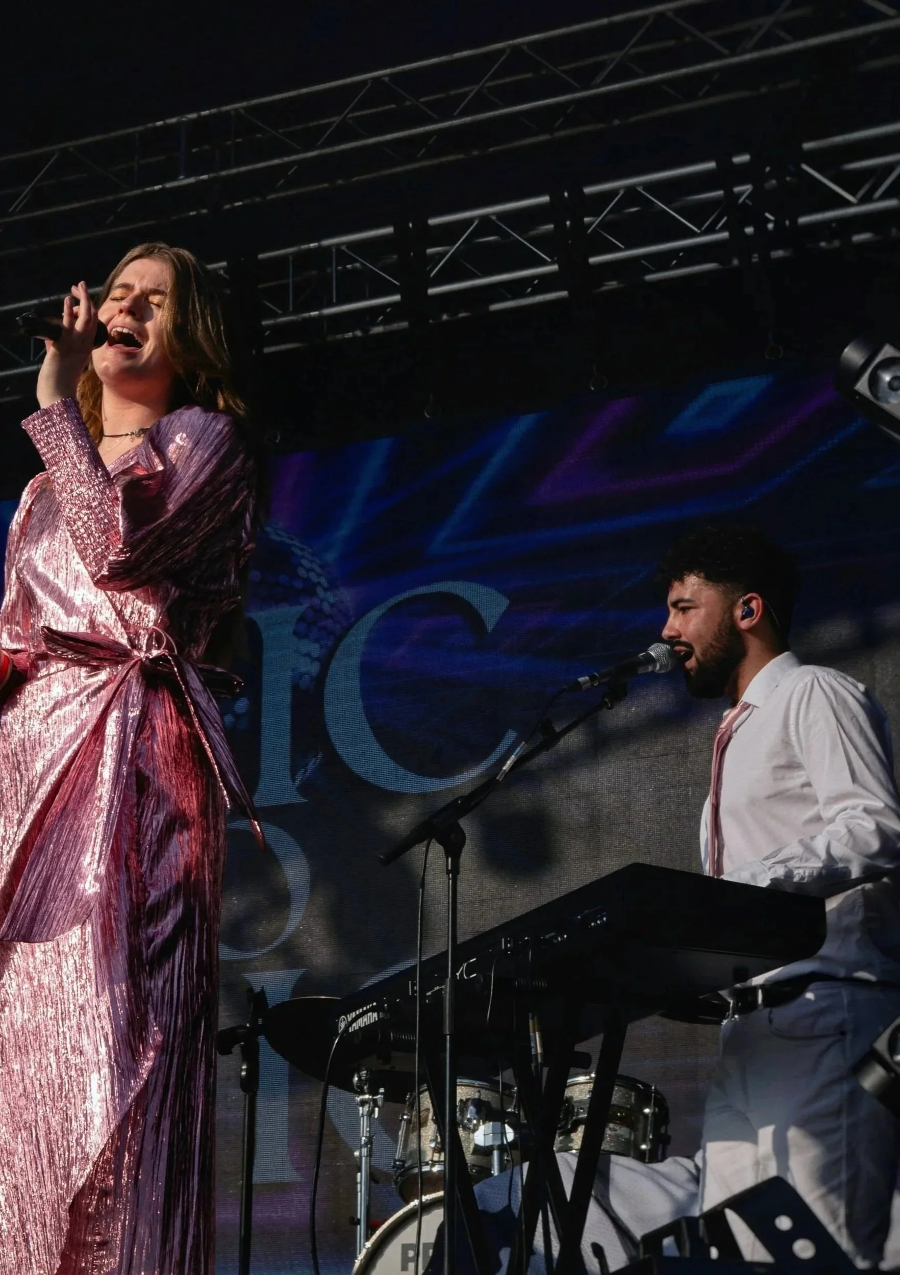 Female singer with brown hair singing into a microphone on stage, wearing a shiny pink dress. Nearby, a male musician with a beard and white shirt is playing an electronic keyboard, with stage lighting and a dark background.
