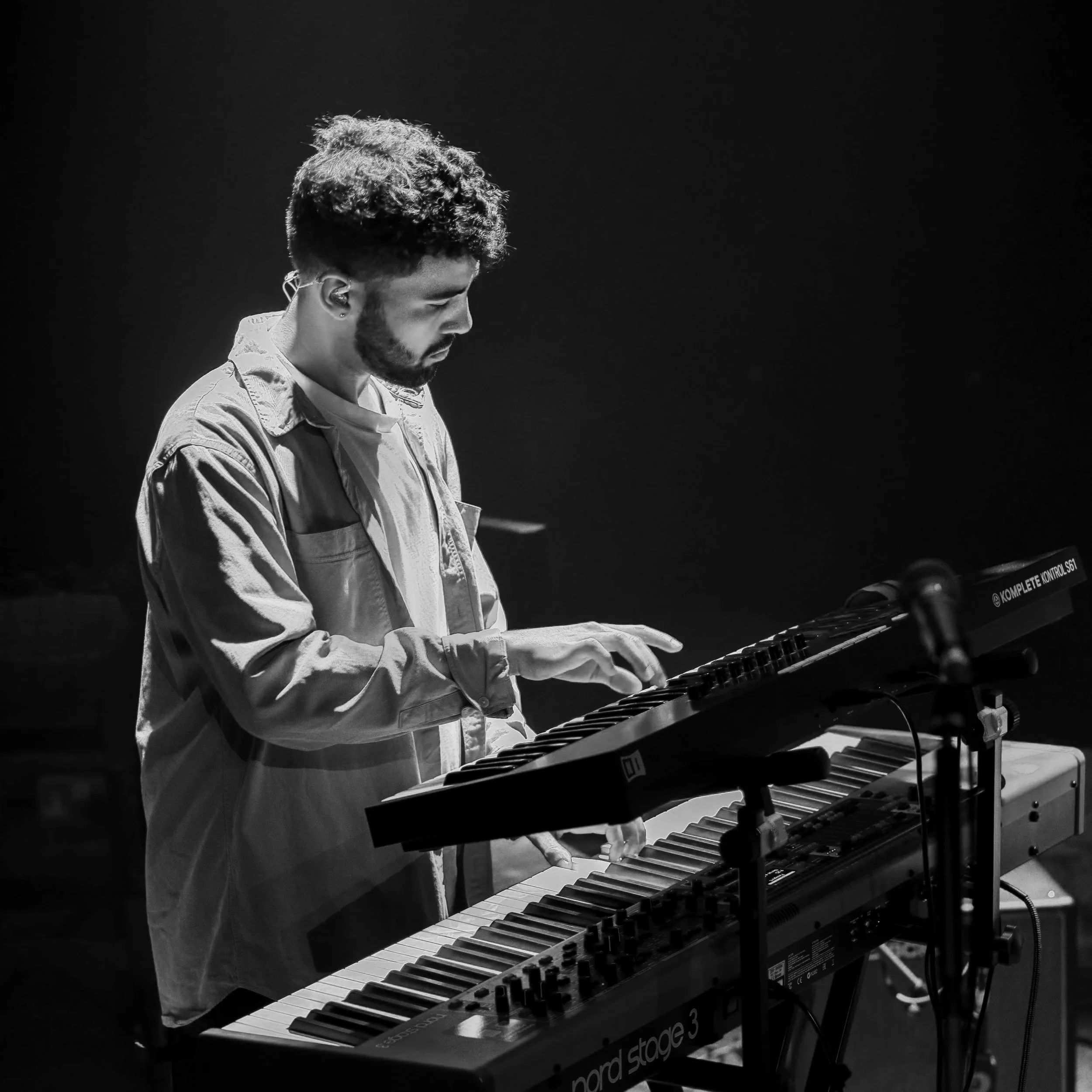 A man with curly hair and a beard playing a keyboard on stage, in a black and white photo with a dark background.