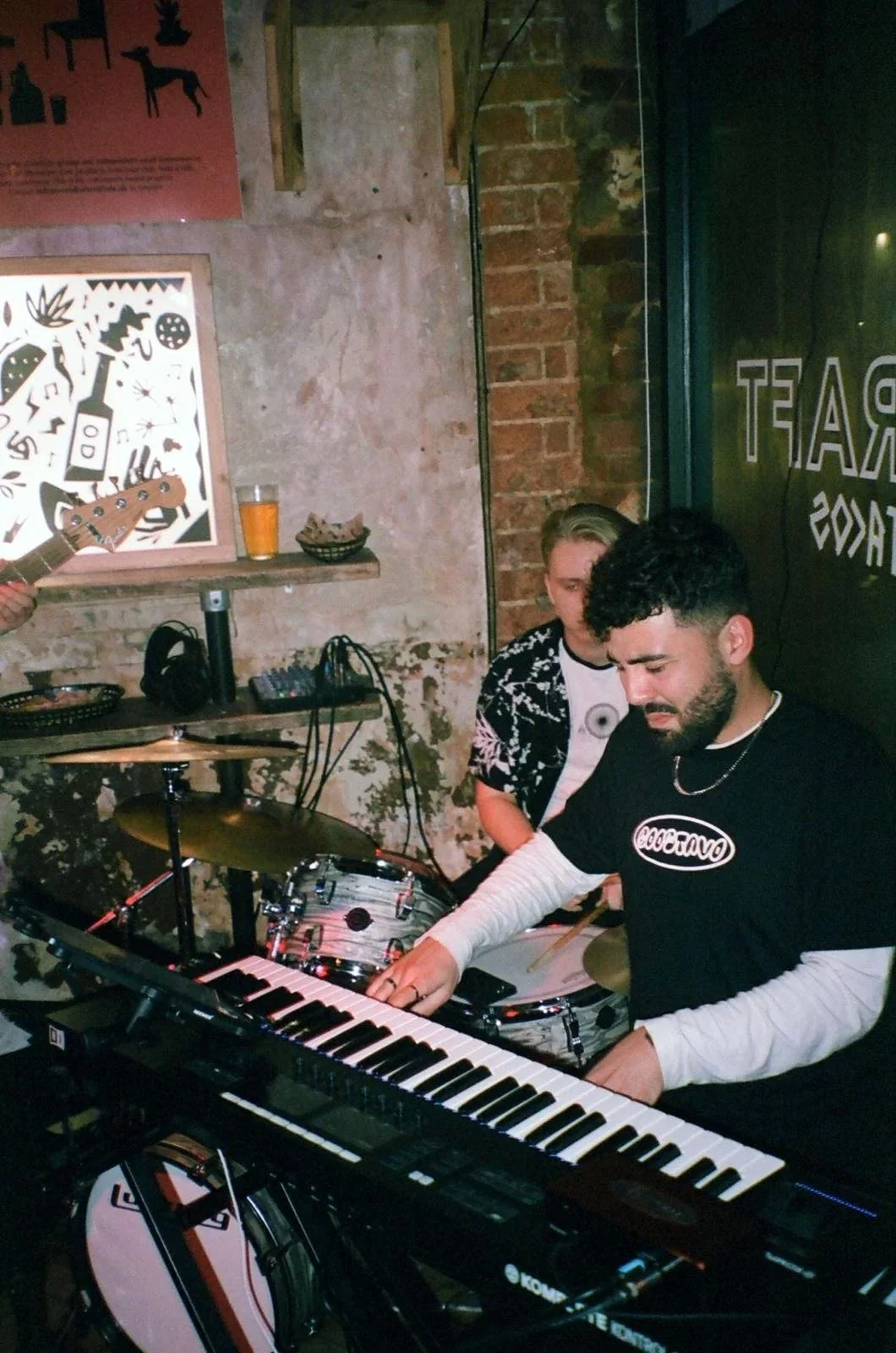 Two men are performing music in a dimly lit venue. One is playing a keyboard, and the other is behind a drum set. Various musical equipment and a drink are visible in the background.