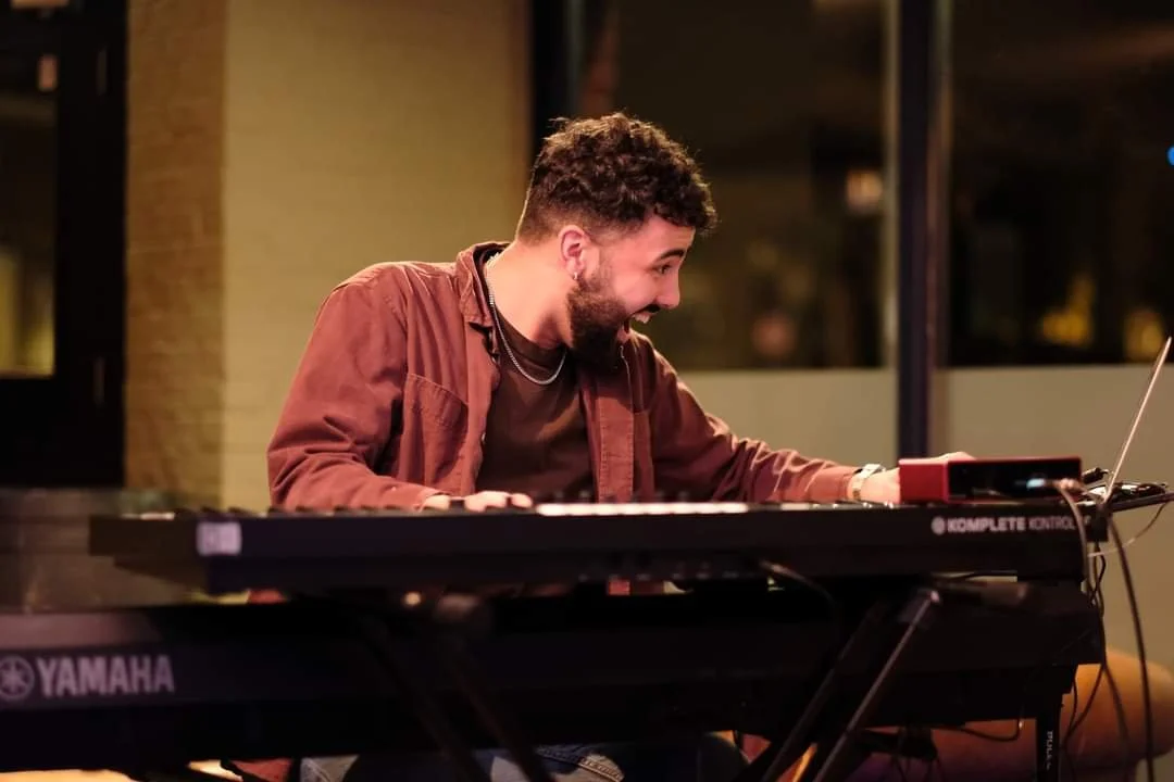 A man with dark curly hair and a beard smiling and playing a keyboard during a performance in an indoor setting.