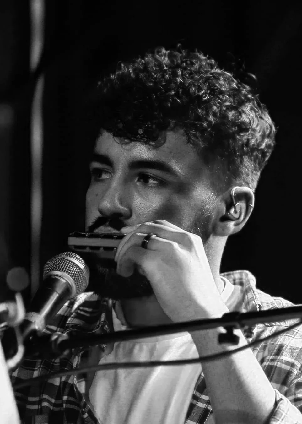 A young man with curly hair playing a harmonica in front of a microphone, with an in-ear monitor in his right ear, in a black and white photo.
