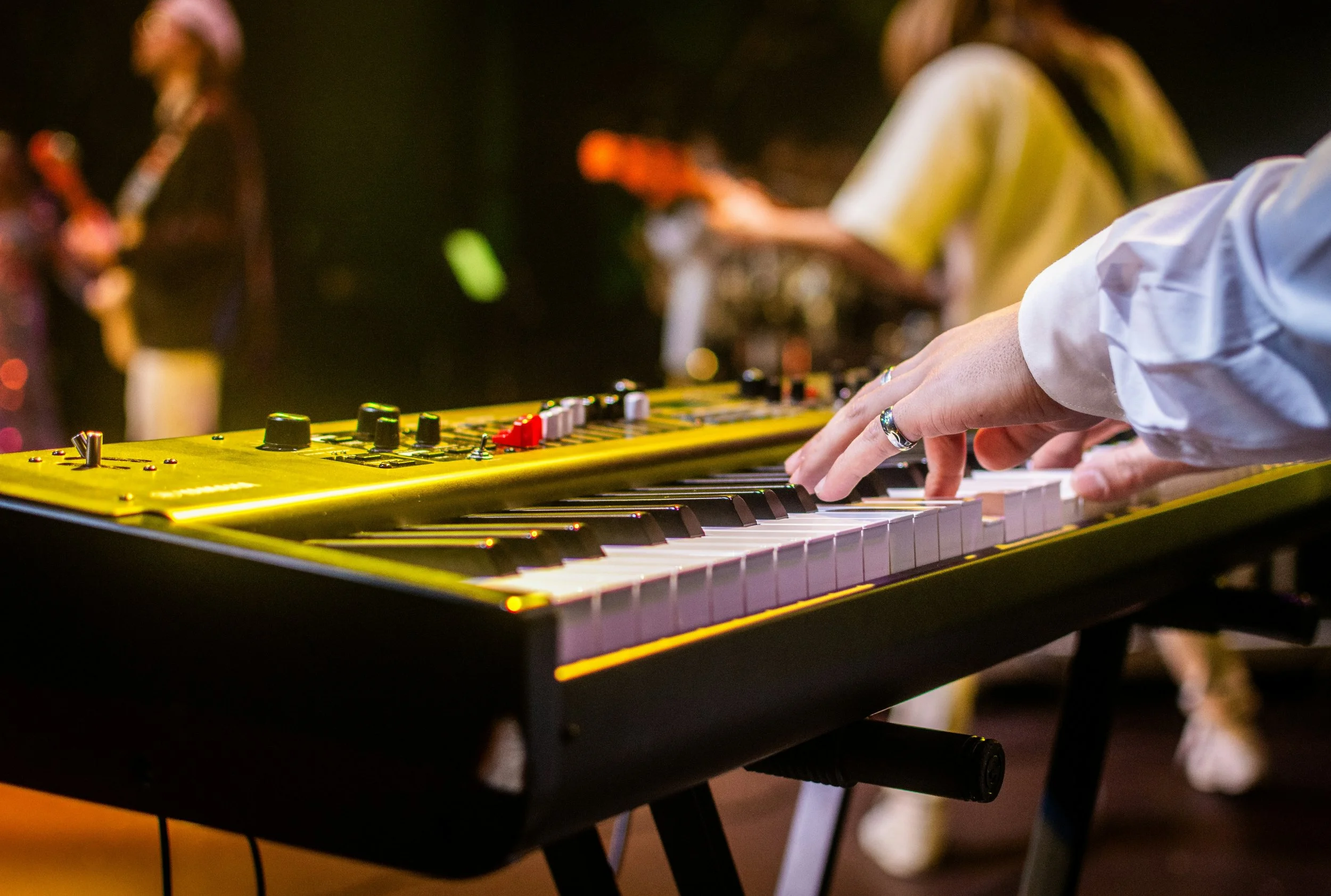 A musician playing a yellow electronic keyboard with a black stand in a dimly-lit setting, with other people in the background, possibly at a live music event or performance.