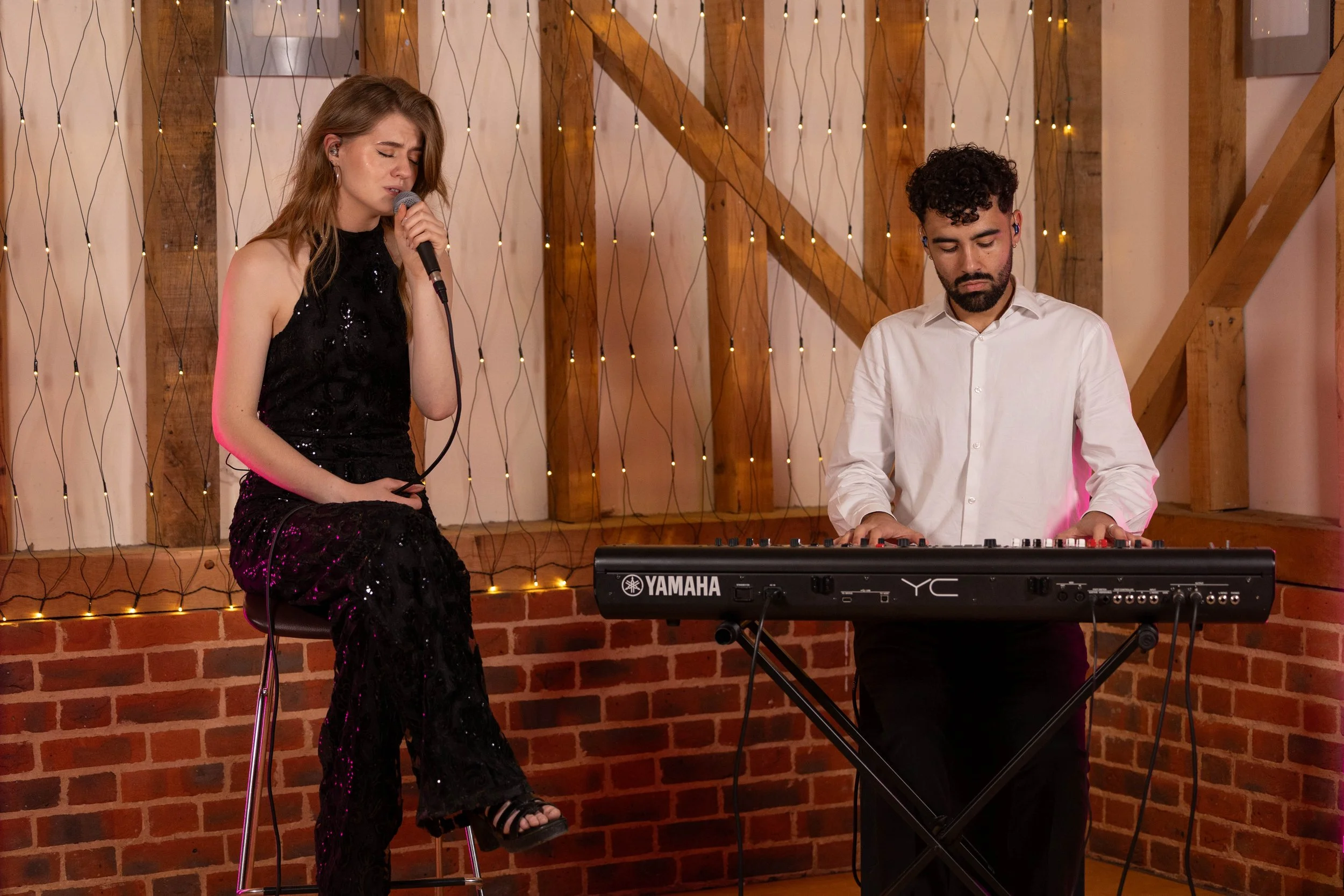 A woman singing with a microphone while a man plays keyboard in a cozy, warmly lit setting with a brick wall and string lights in the background.