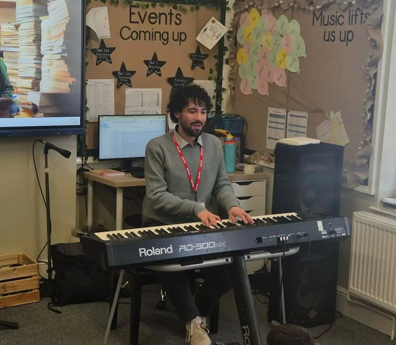 A man playing a Roland RD-300NX digital piano in a classroom or music room, with computer monitors and educational posters on the wall behind him.