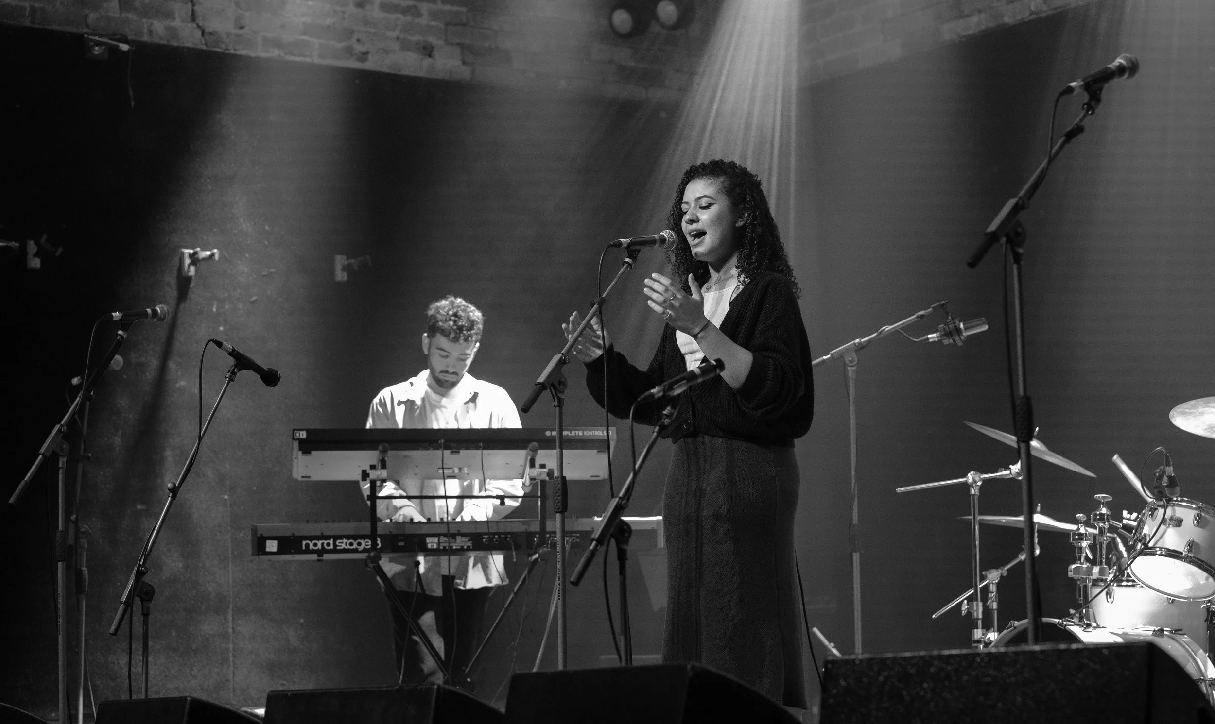 A black and white photo of a musical performance on stage. A woman with curly hair sings into a microphone, while a man plays a keyboard behind her. There are multiple microphones and musical instrument stands around them, and stage lighting creates spotlights on the performers.