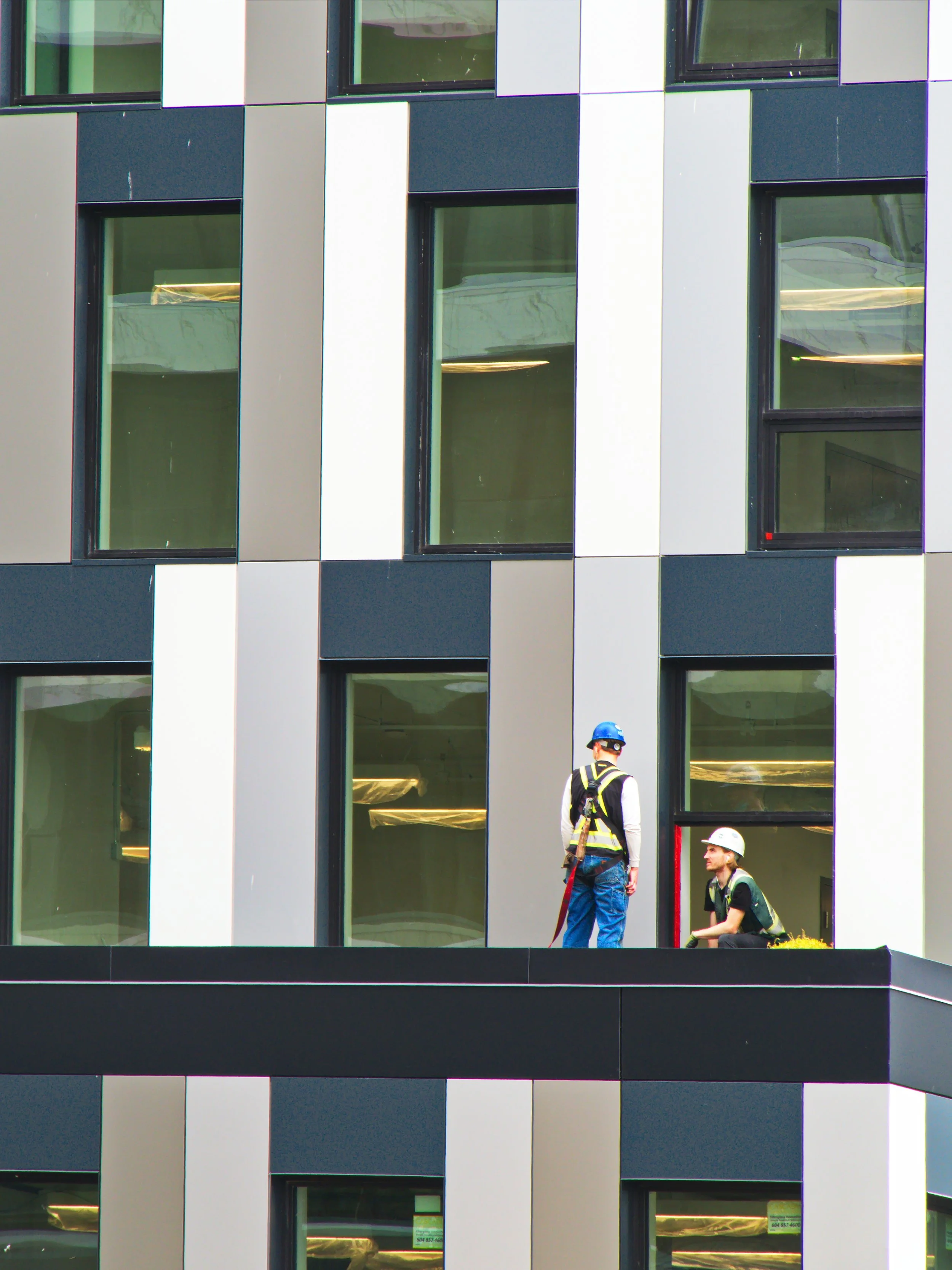 Two construction workers wearing helmets on a balcony of a modern building with large windows.