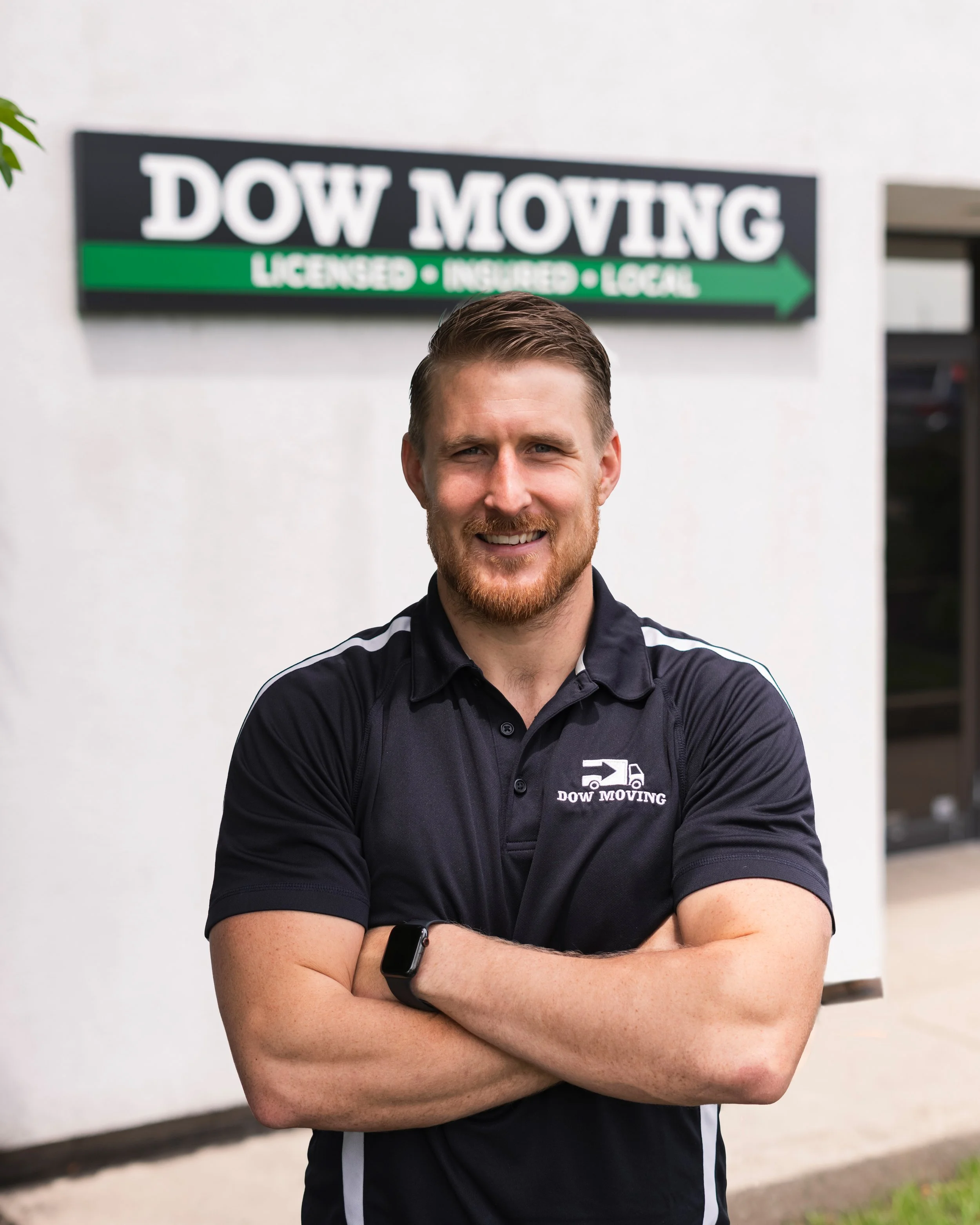 A man with a beard smiling, wearing a black polo shirt with a logo, standing in front of a white wall with a sign that says 'DOW MOVING' and 'LICENSED, INSURED, LOCAL'.