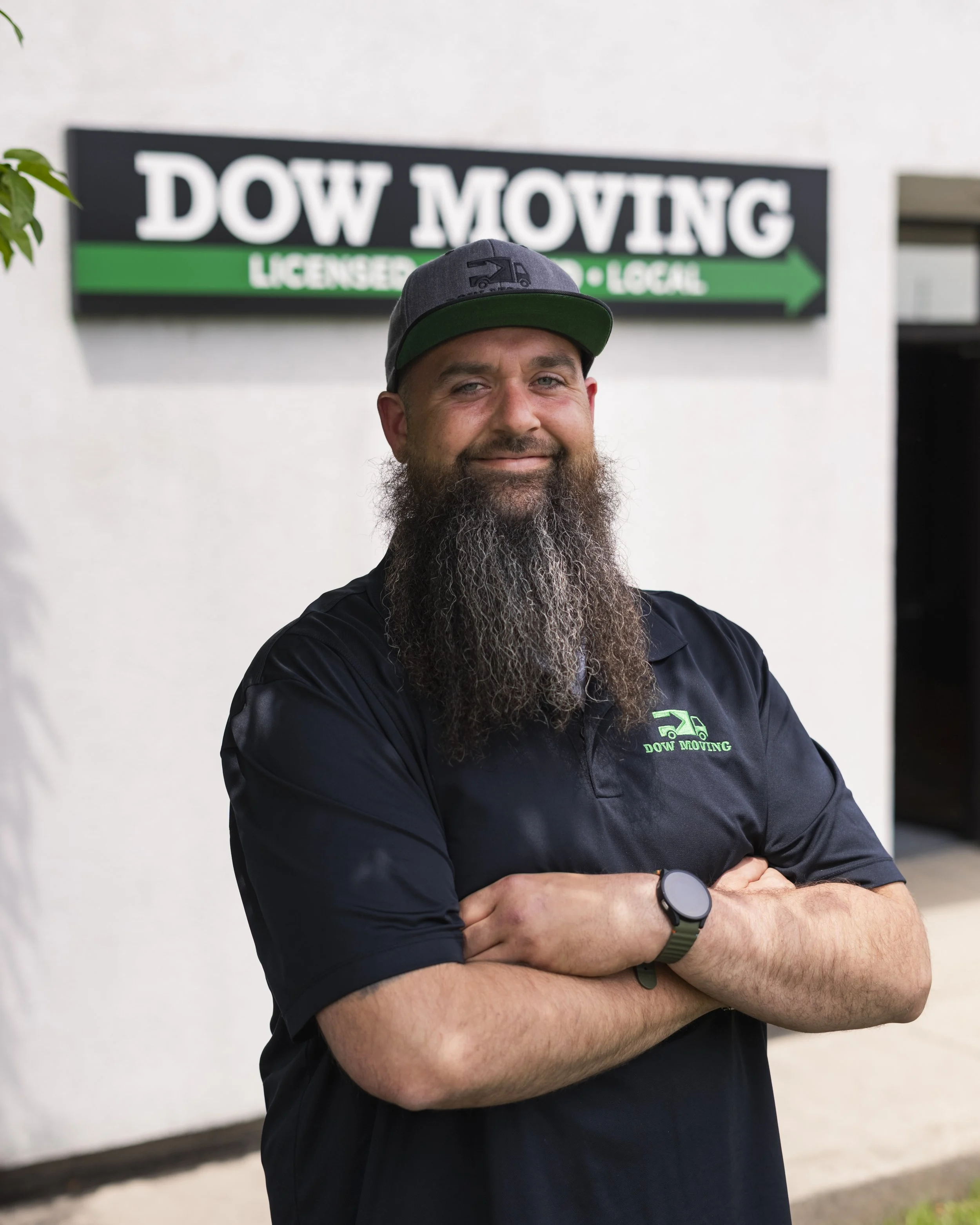 A man with a beard and a gray cap standing in front of a sign that says 'DOW MOVING' with a green arrow and logo, wearing a black polo shirt with the company's logo and a smartwatch, crossing his arms and smiling.