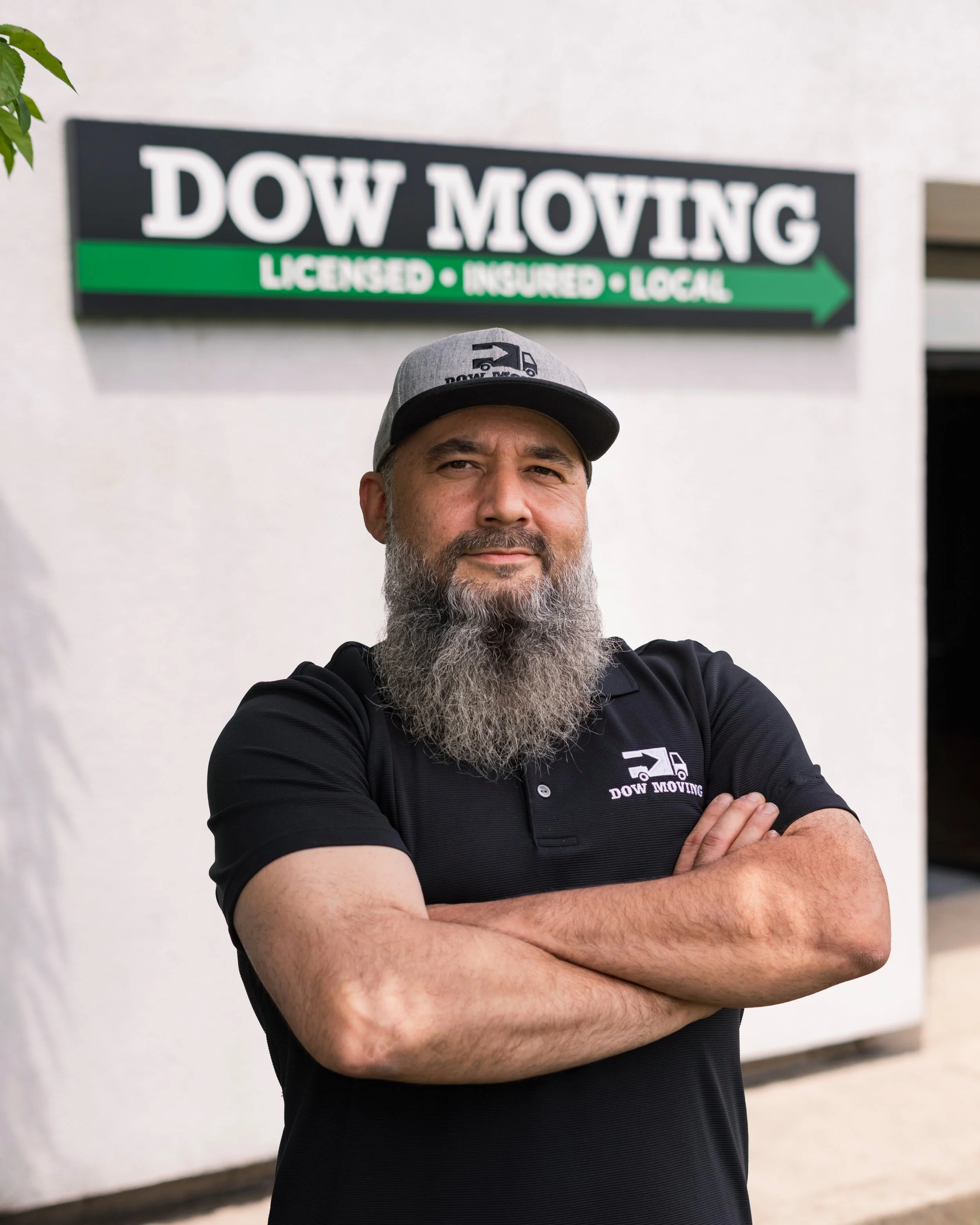 A man with a gray beard and mustache standing with arms crossed, wearing a black polo shirt and a gray cap with a Dow Moving logo, in front of a white wall with a sign for Dow Moving, a moving company.