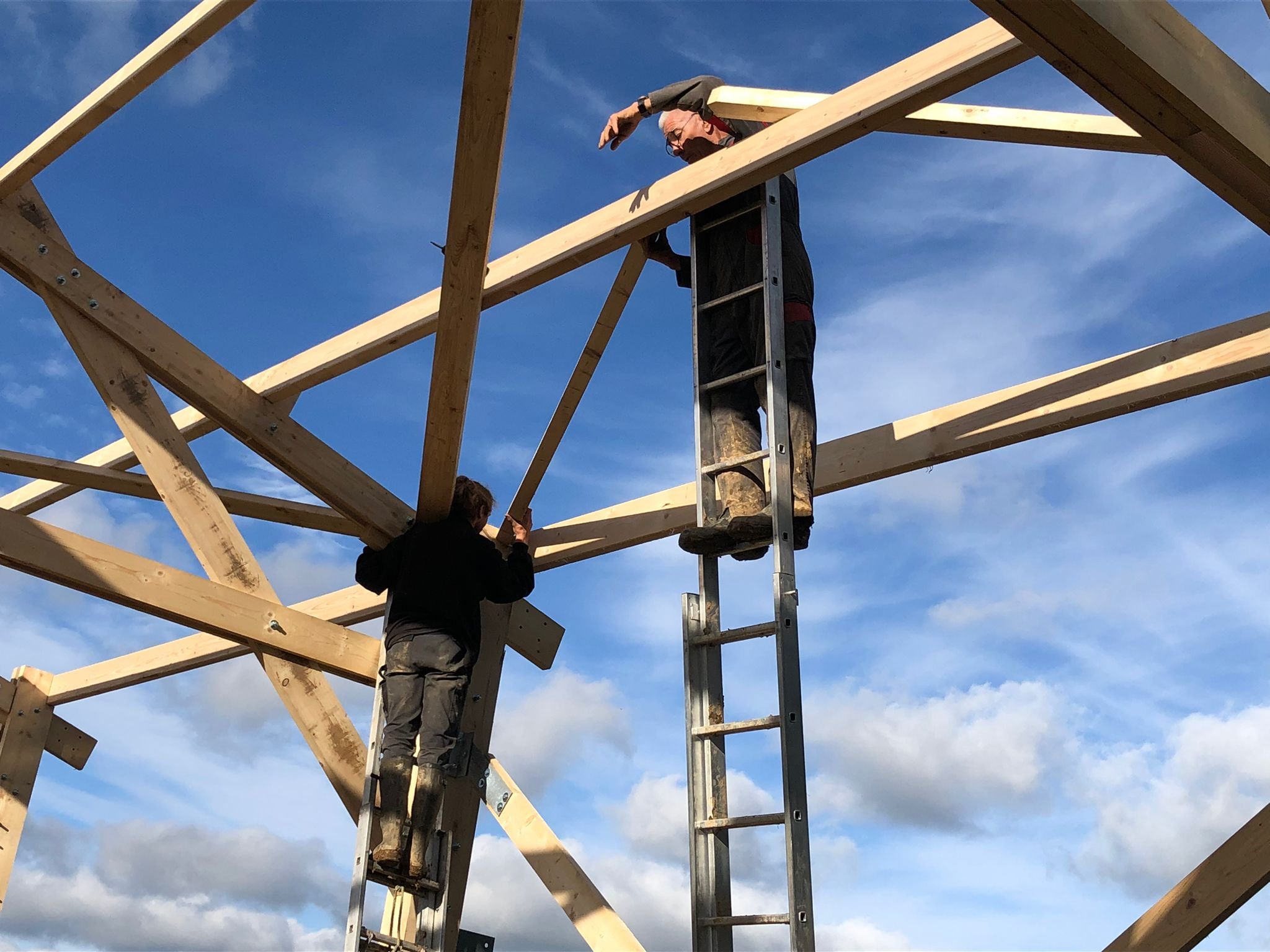 Deux personnes construisent une structure en bois, l'une en haut sur une échelle et l'autre en bas, contre un ciel bleu avec quelques nuages.