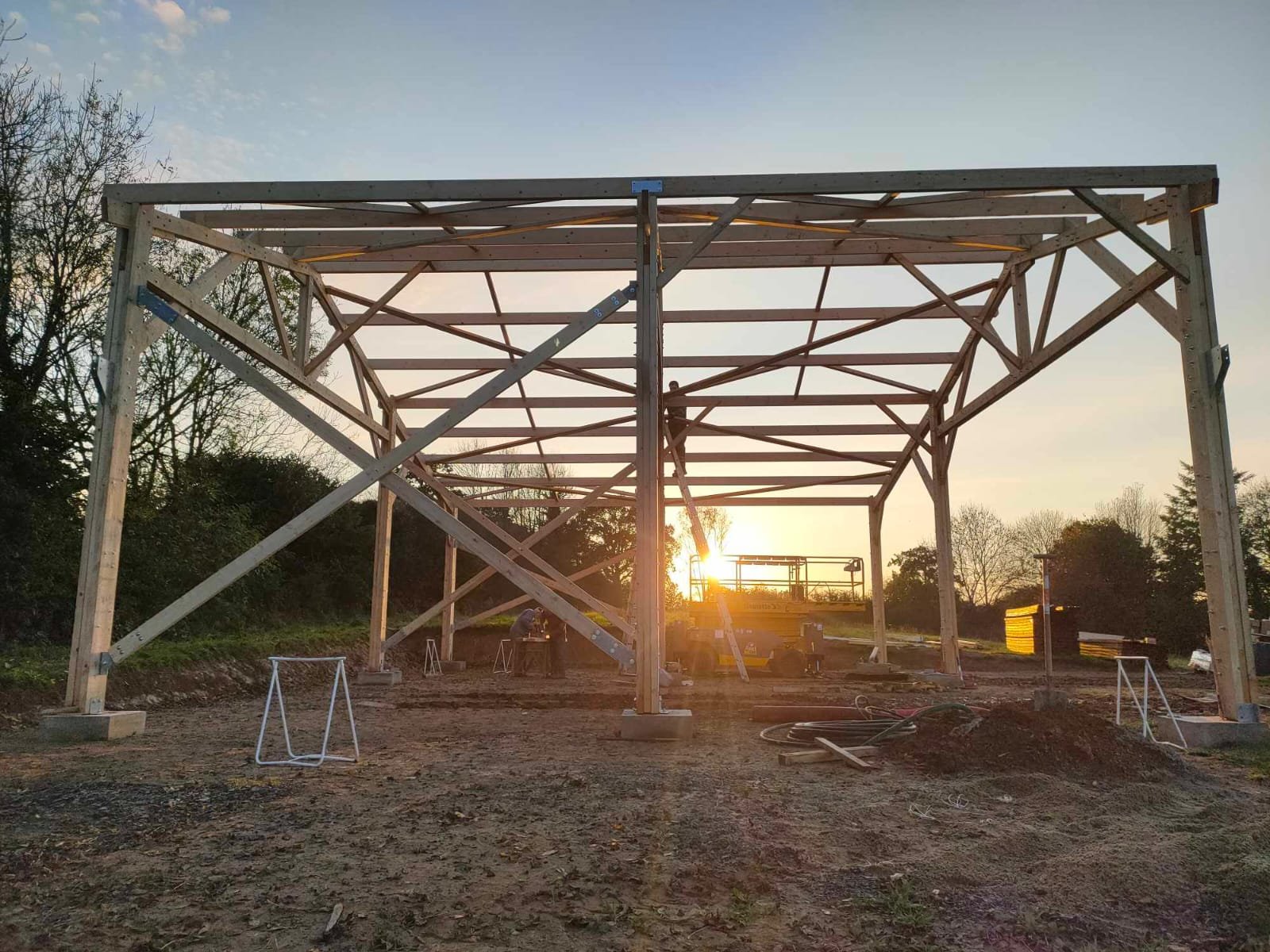 Une structure en bois en cours de construction, avec des outils et des matériaux sur le sol, au coucher du soleil.